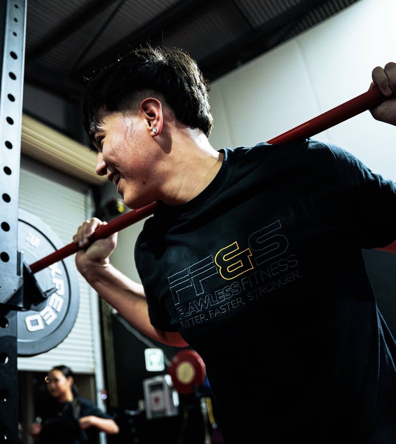 A woman with short dark hair and earrings doing a squat with a red barbell across her shoulders in a gym, smiling and sweating.