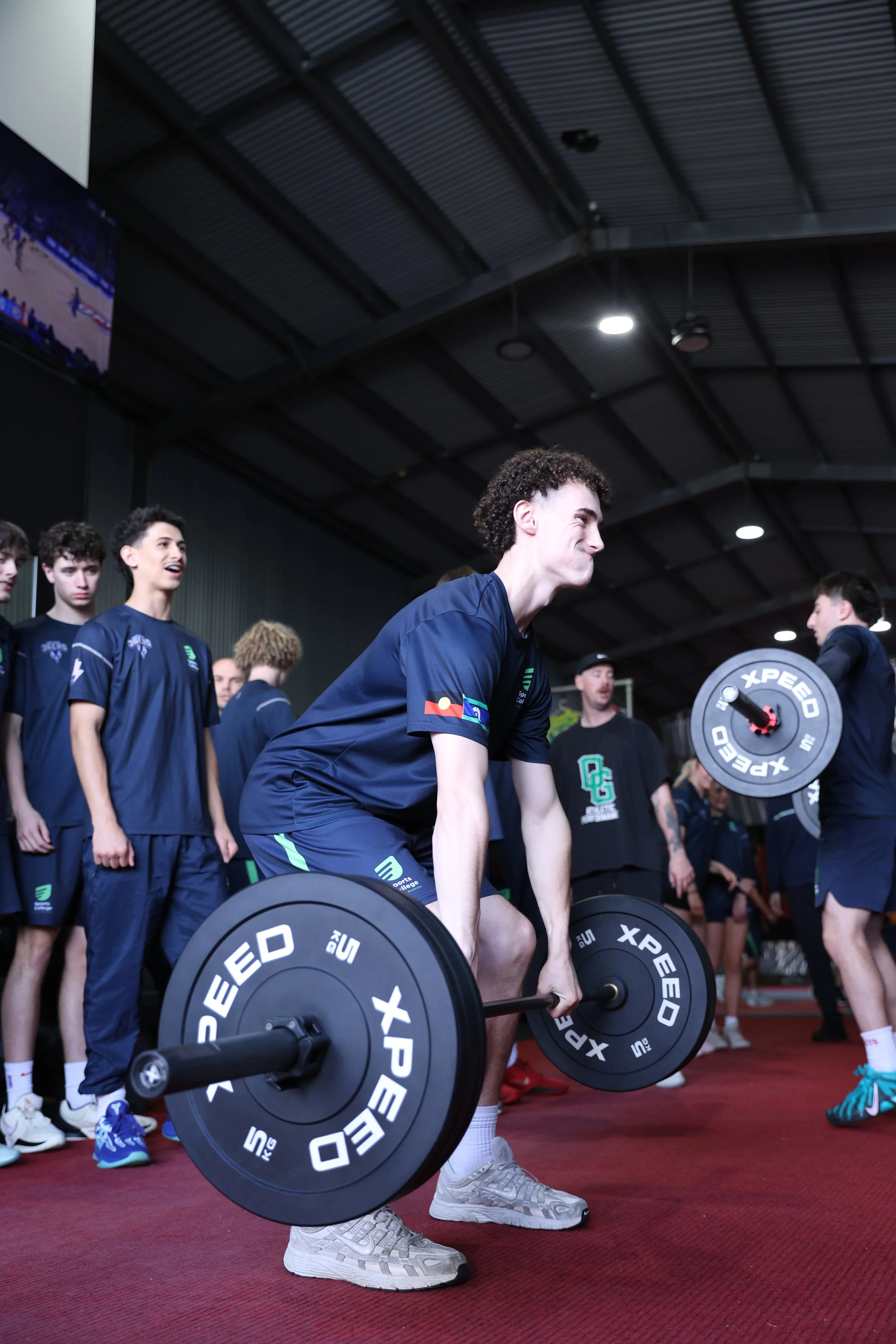 Young man in athletic wear lifting a barbell during a weights training or workout session with a group of people in the background.