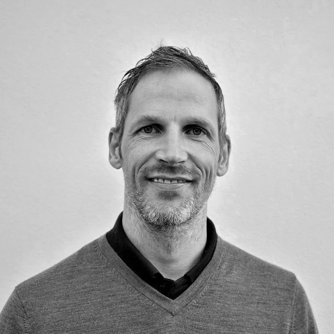 Black and white portrait of a man with short hair, a beard, wearing a collared shirt and a v-neck sweater, smiling, standing against a plain wall.