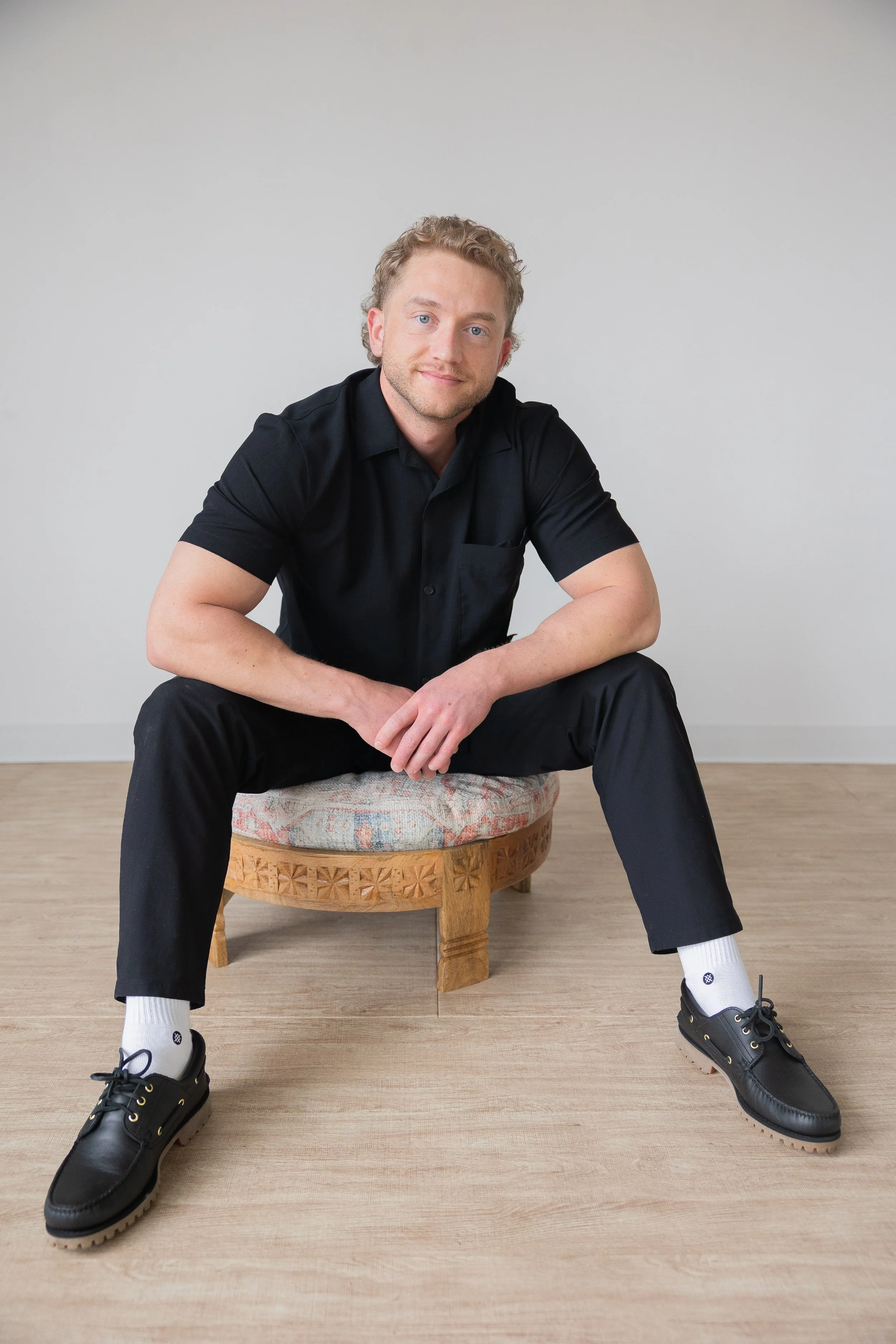 A smiling man sitting on a wooden stool against a white brick wall, wearing a beige t-shirt and dark pants.