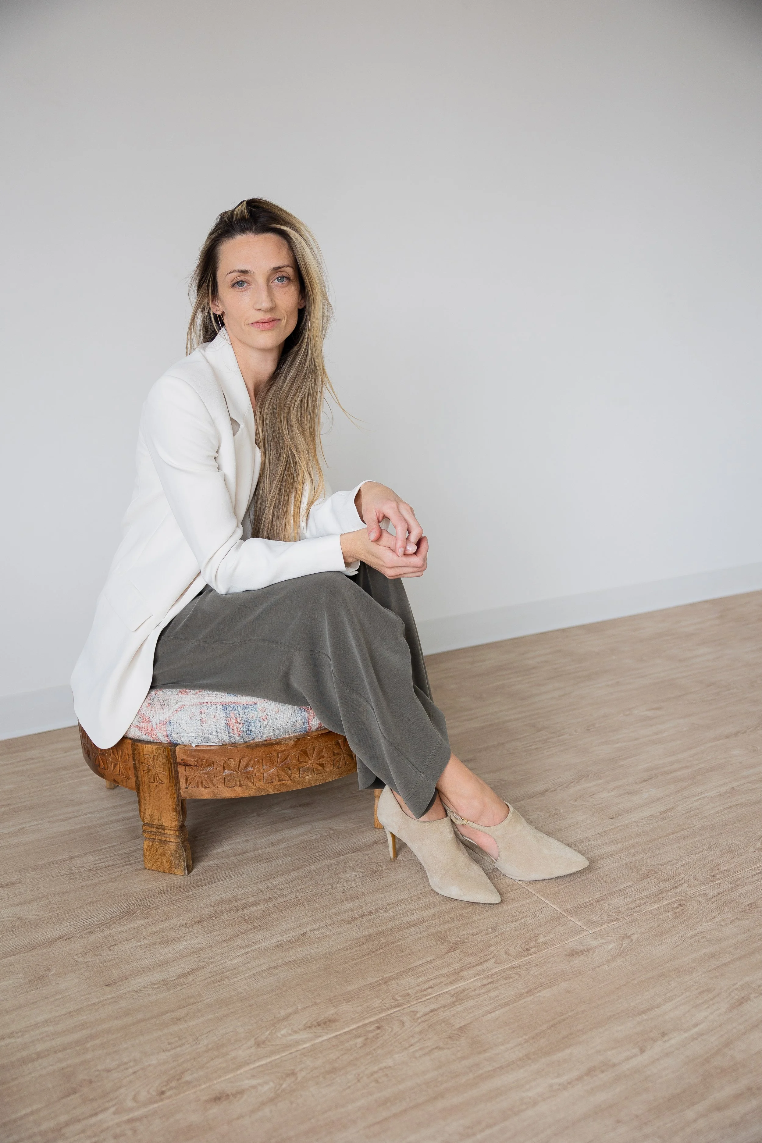 A woman with long wavy hair sitting on a wooden stool in front of a light gray brick wall, wearing a black sleeveless top and white pants.