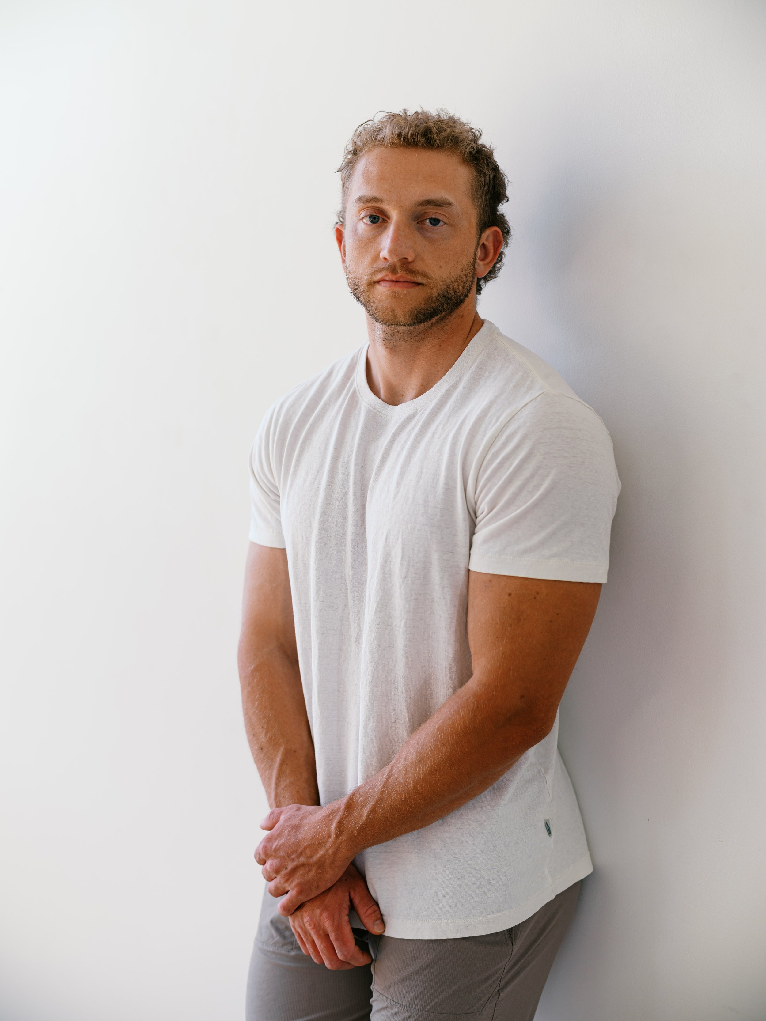 A smiling man sitting on a wooden stool against a white brick wall, wearing a beige t-shirt and dark pants.