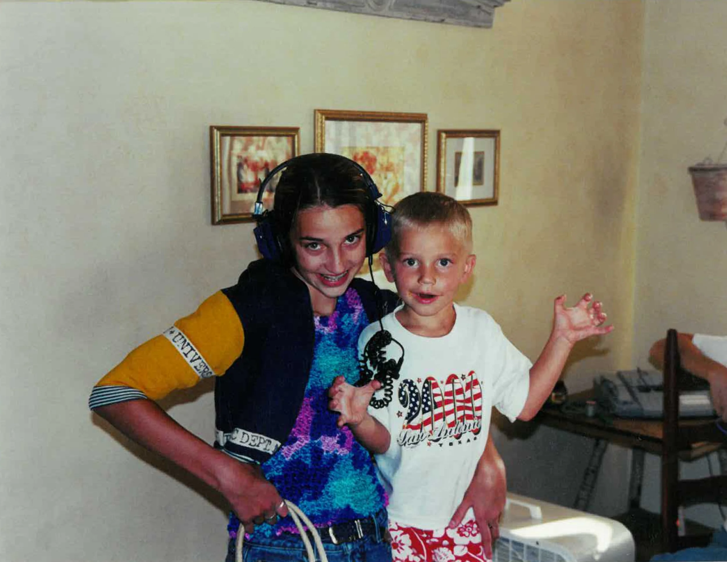 A girl with dark hair wearing large headphones and a yellow and black shirt, standing next to a young boy with short blonde hair wearing a white T-shirt and red patterned shorts, both smiling and looking at the camera in a room with framed artwork and a table with a telephone and other items.