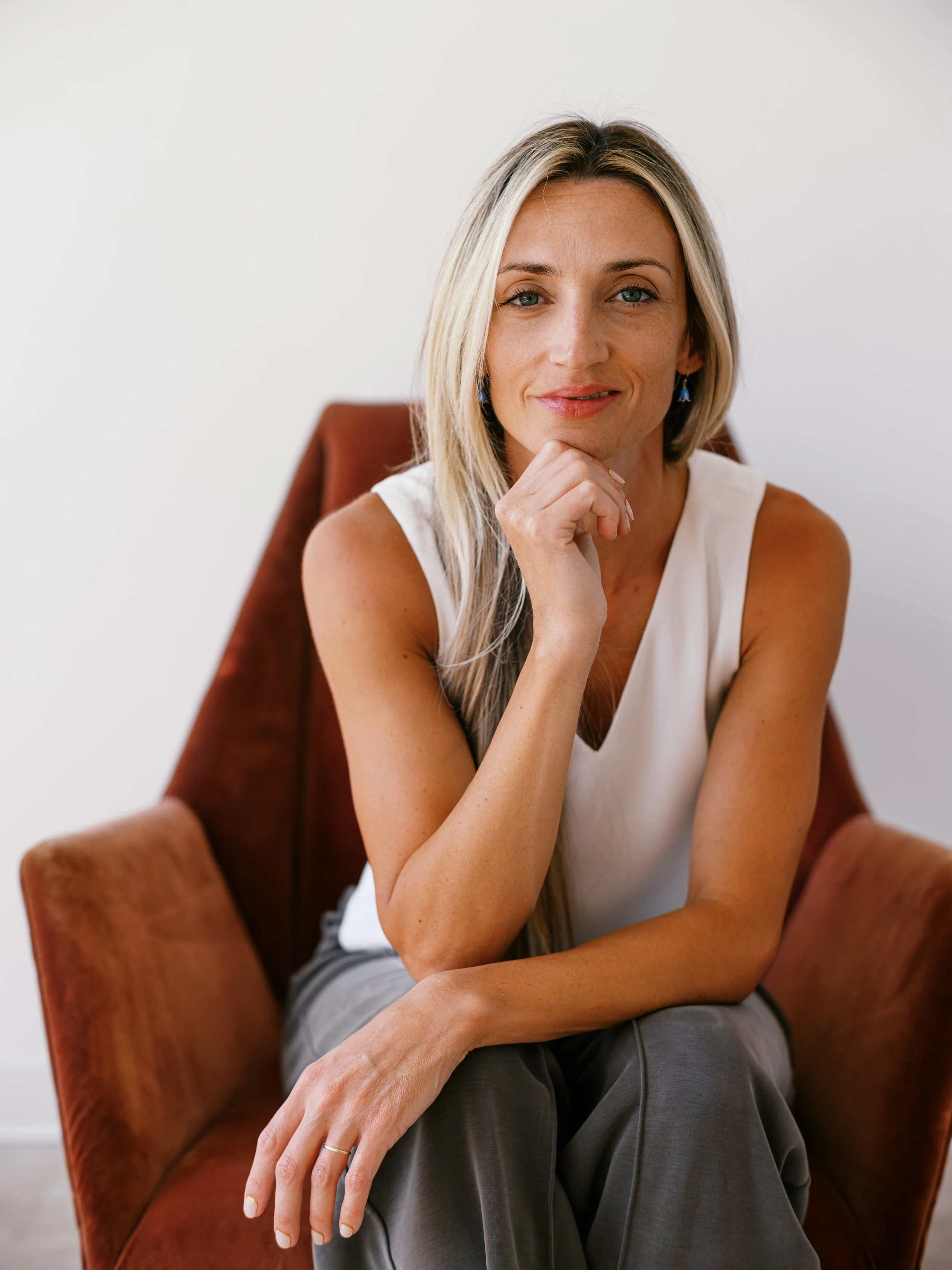 A woman with long wavy hair sitting on a wooden stool in front of a light gray brick wall, wearing a black sleeveless top and white pants.