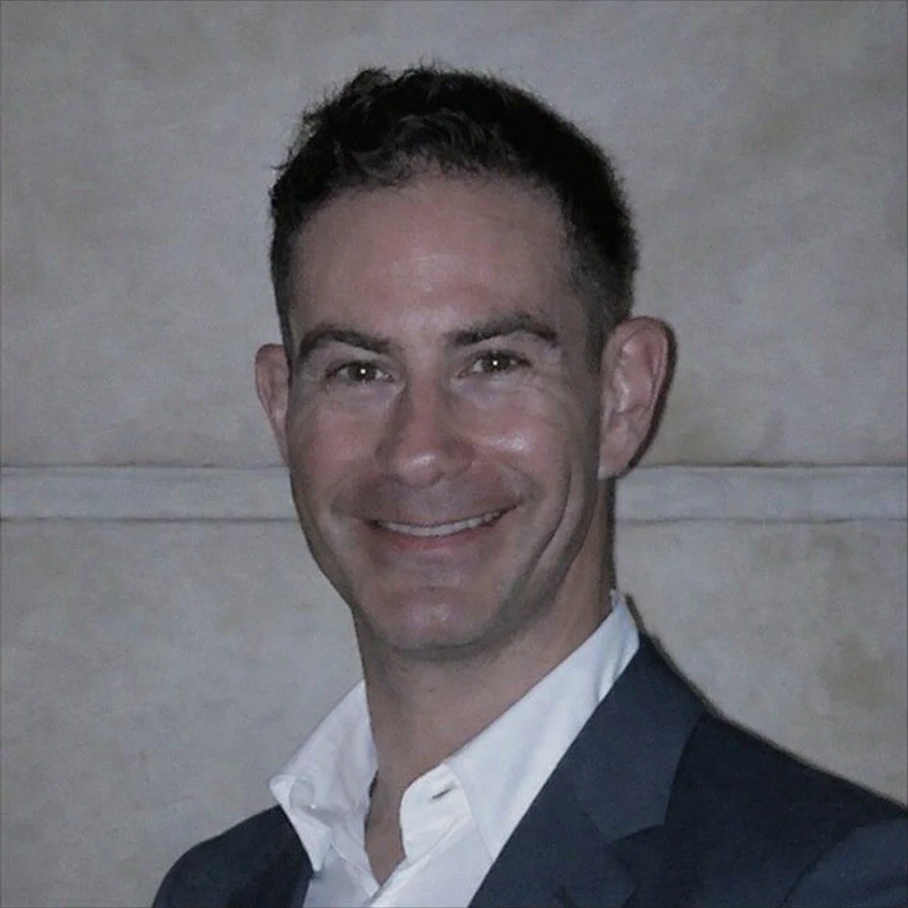 A smiling man with dark hair wearing a white shirt and dark suit in front of a light-colored wall.