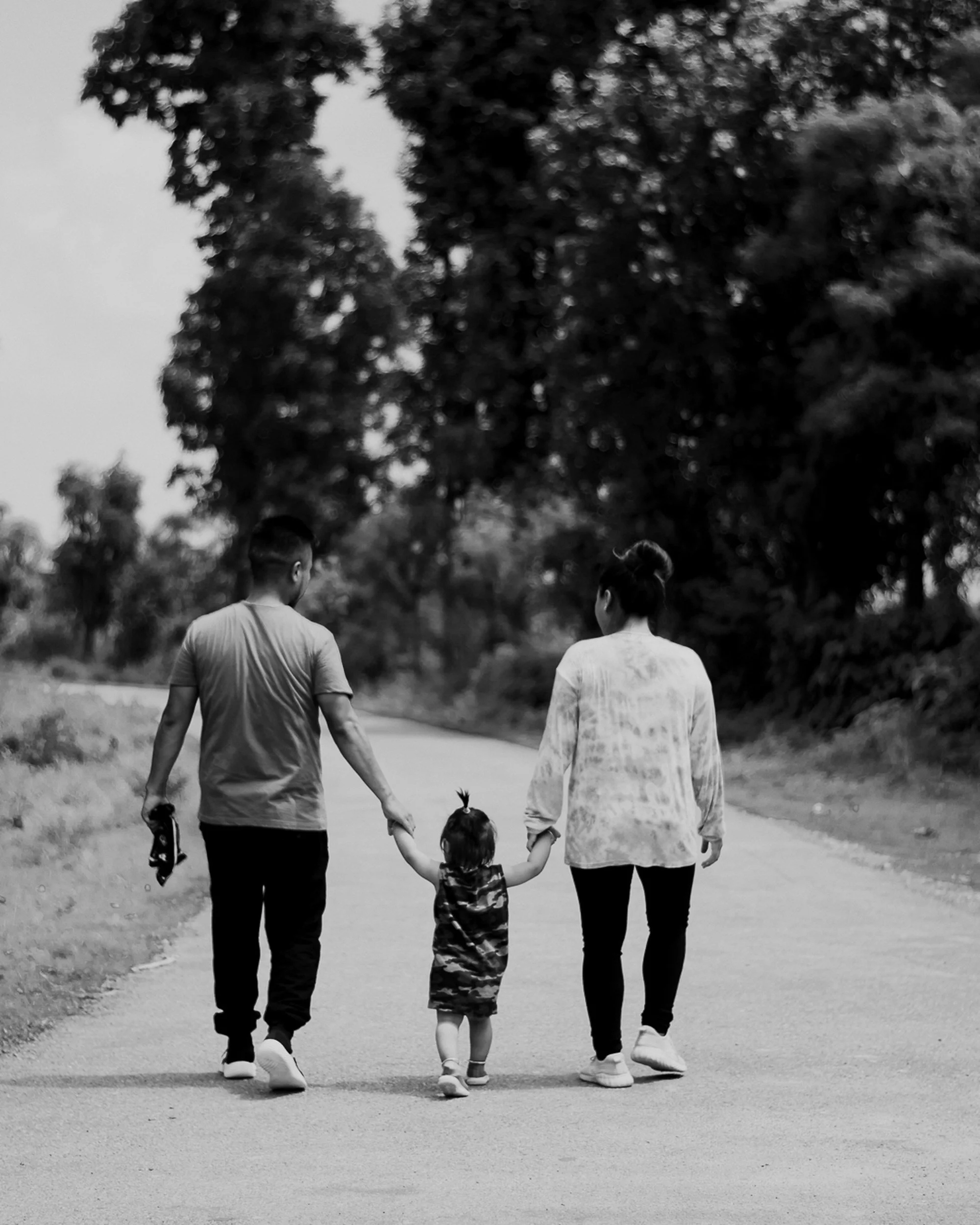 A family of four walking hand-in-hand on a dirt path through a park with trees in the background. The photo is in black and white.