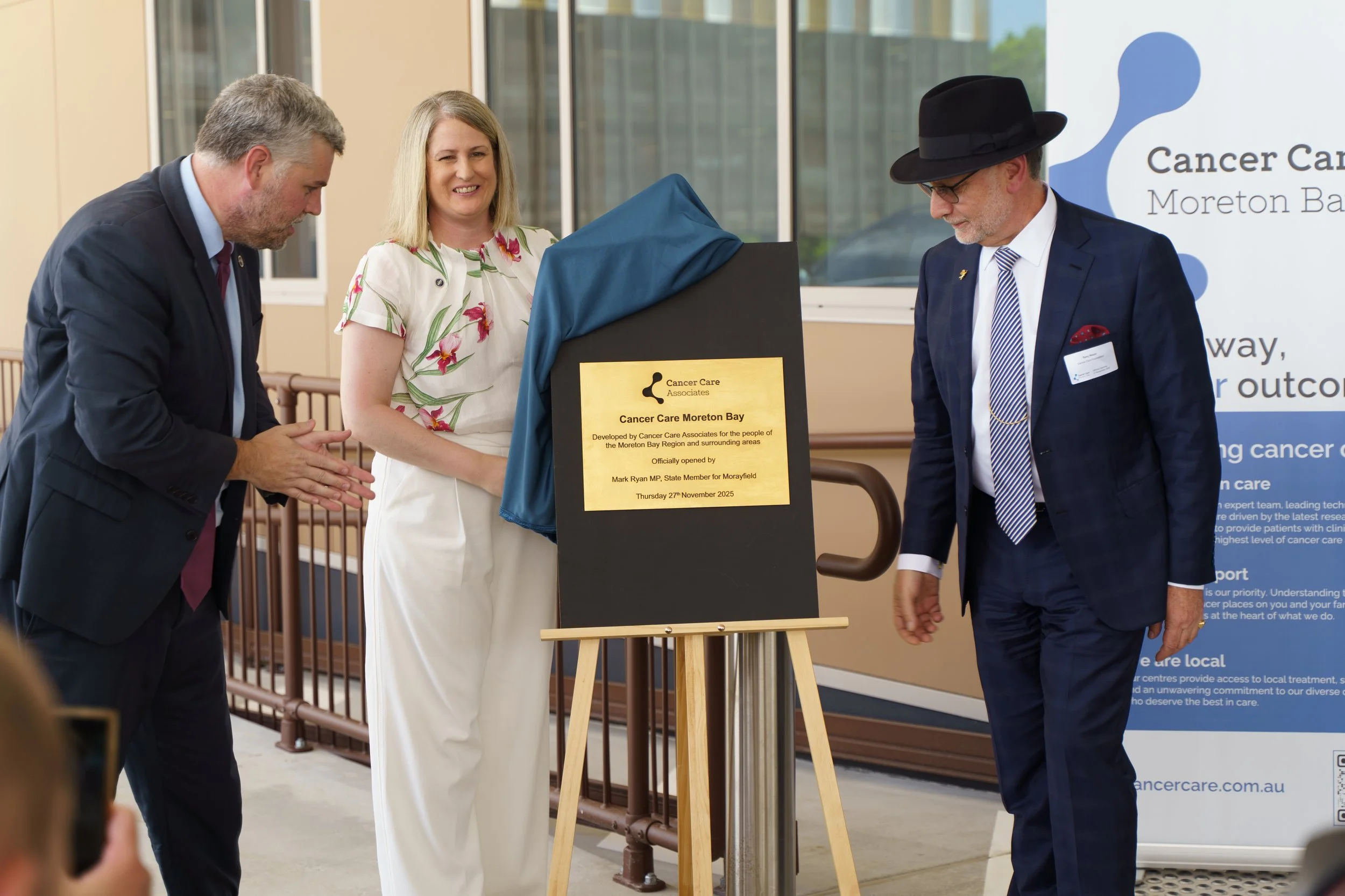 Three people standing around a plaque at an unveiling ceremony for Cancer Care Moreton Bay. The man on the left is clapping, the woman in the middle is smiling, and the man on the right is wearing a black hat and suit. The plaque is covered with a bl