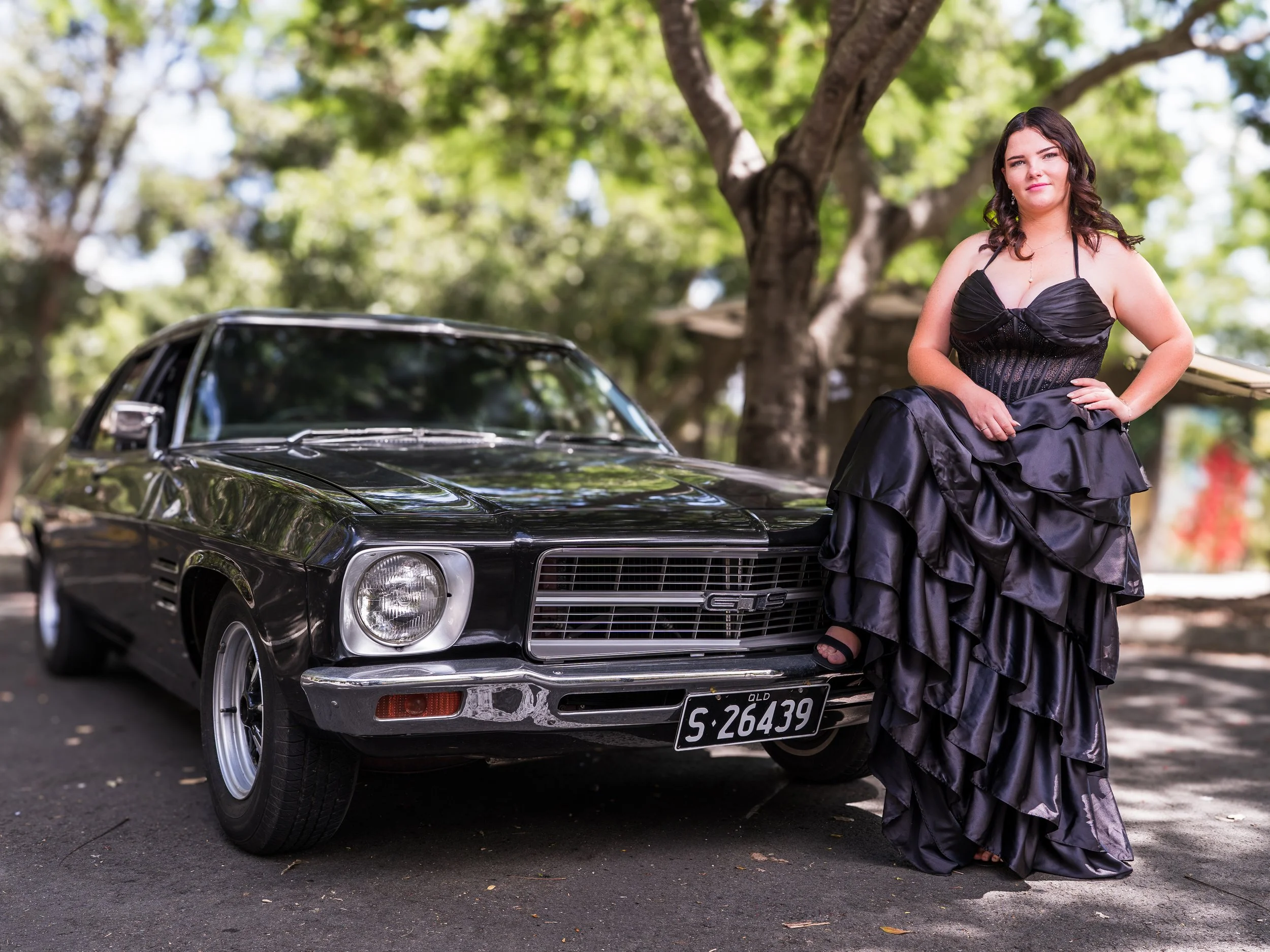 A woman in a black evening gown leaning on a vintage black Camaro car outdoors with trees in the background.