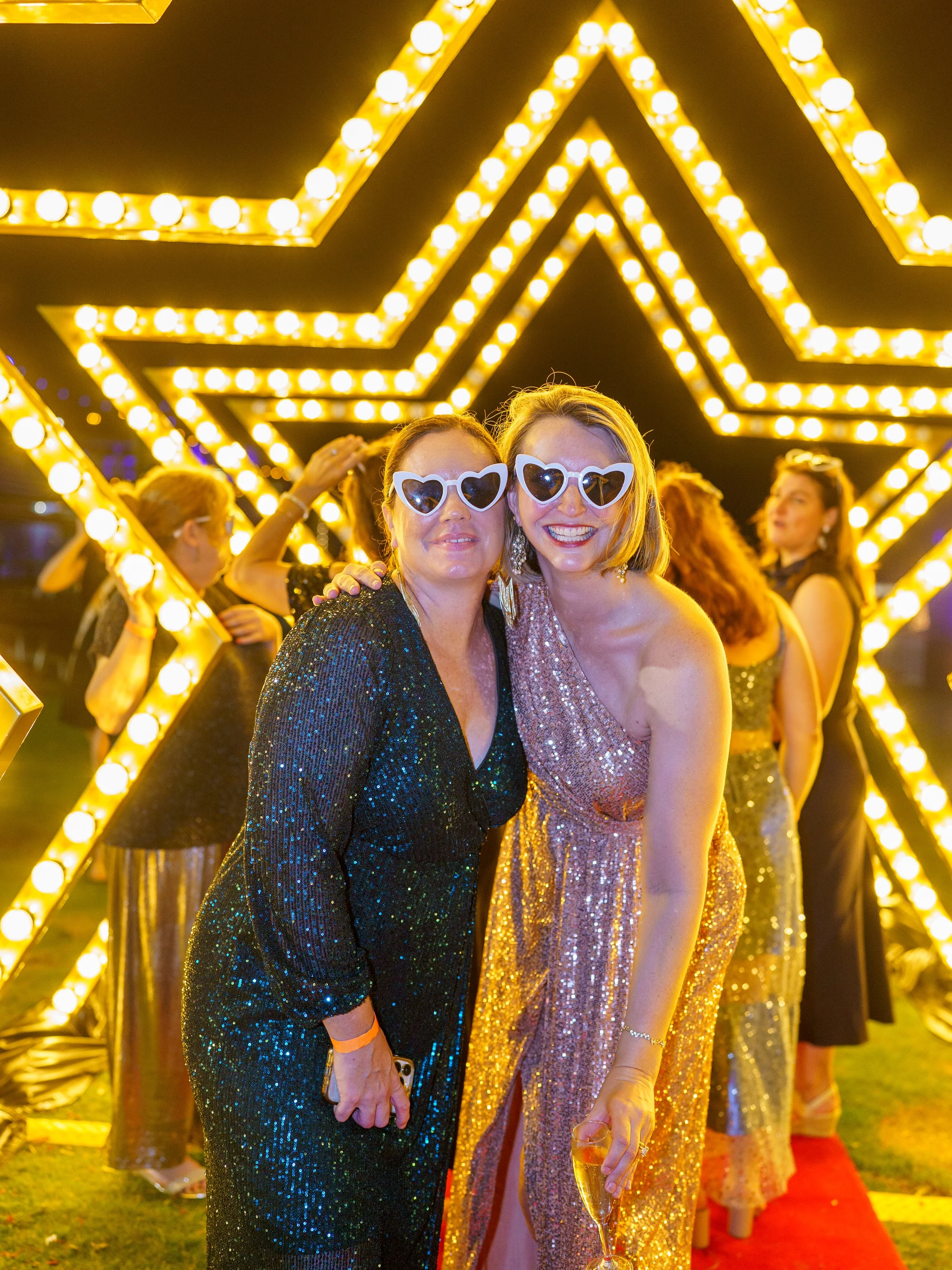 Two women in sequined dresses and heart-shaped sunglasses smiling at a nighttime event with a large illuminated star in the background.