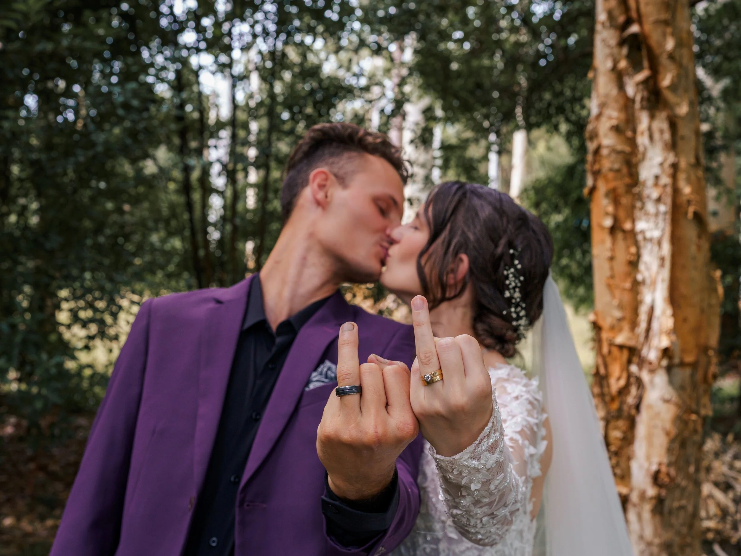 A couple is kissing outdoors in a wooded area. The man is wearing a purple suit jacket and the woman is wearing a white lace dress with a floral headpiece. Their hands are intertwined, showing rings, and the background features trees and blurred sunlight.