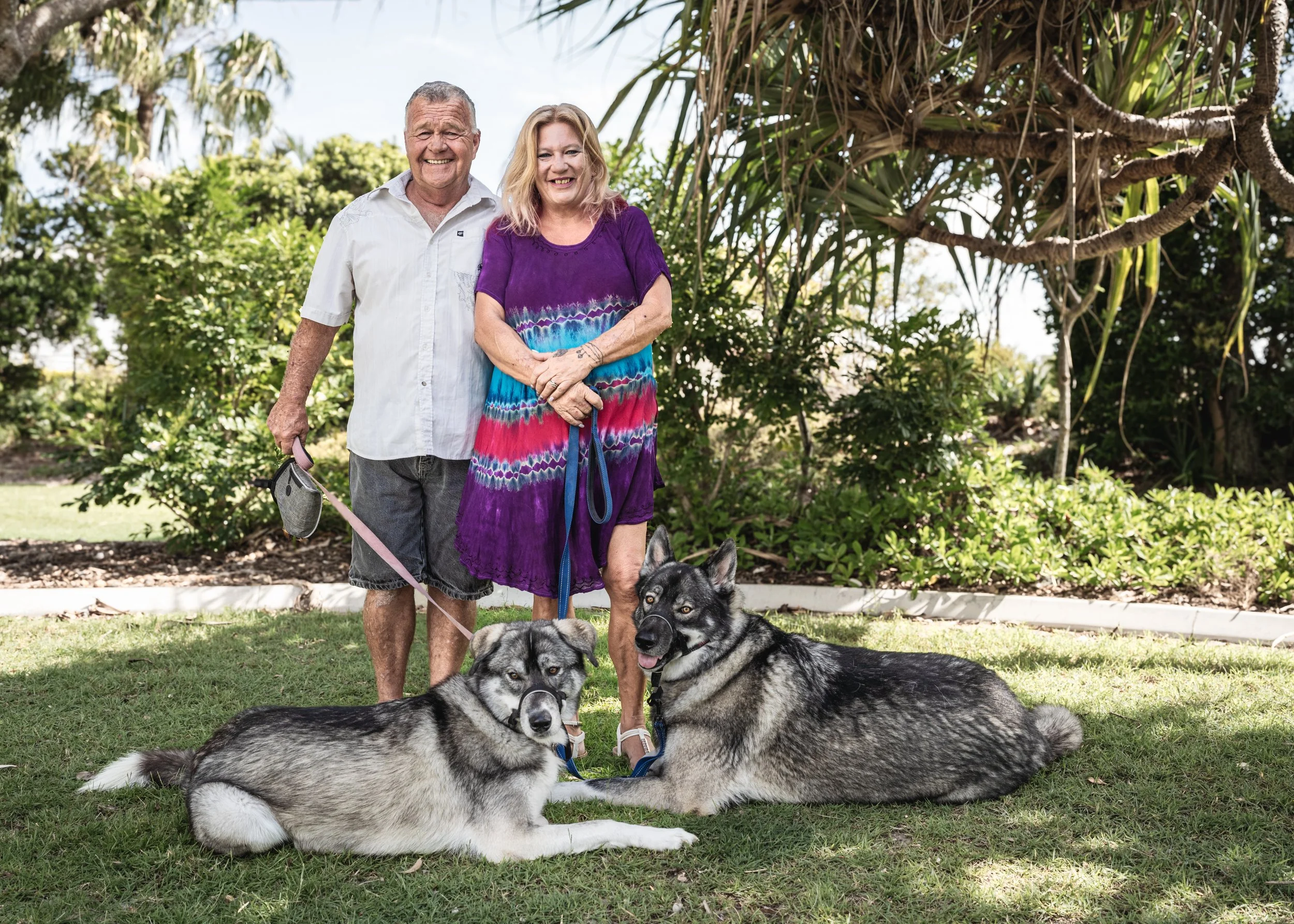 A smiling elderly couple standing outdoors on a sunny day, with two Siberian Huskies lying on the grass in front of them, surrounded by lush trees and bushes.