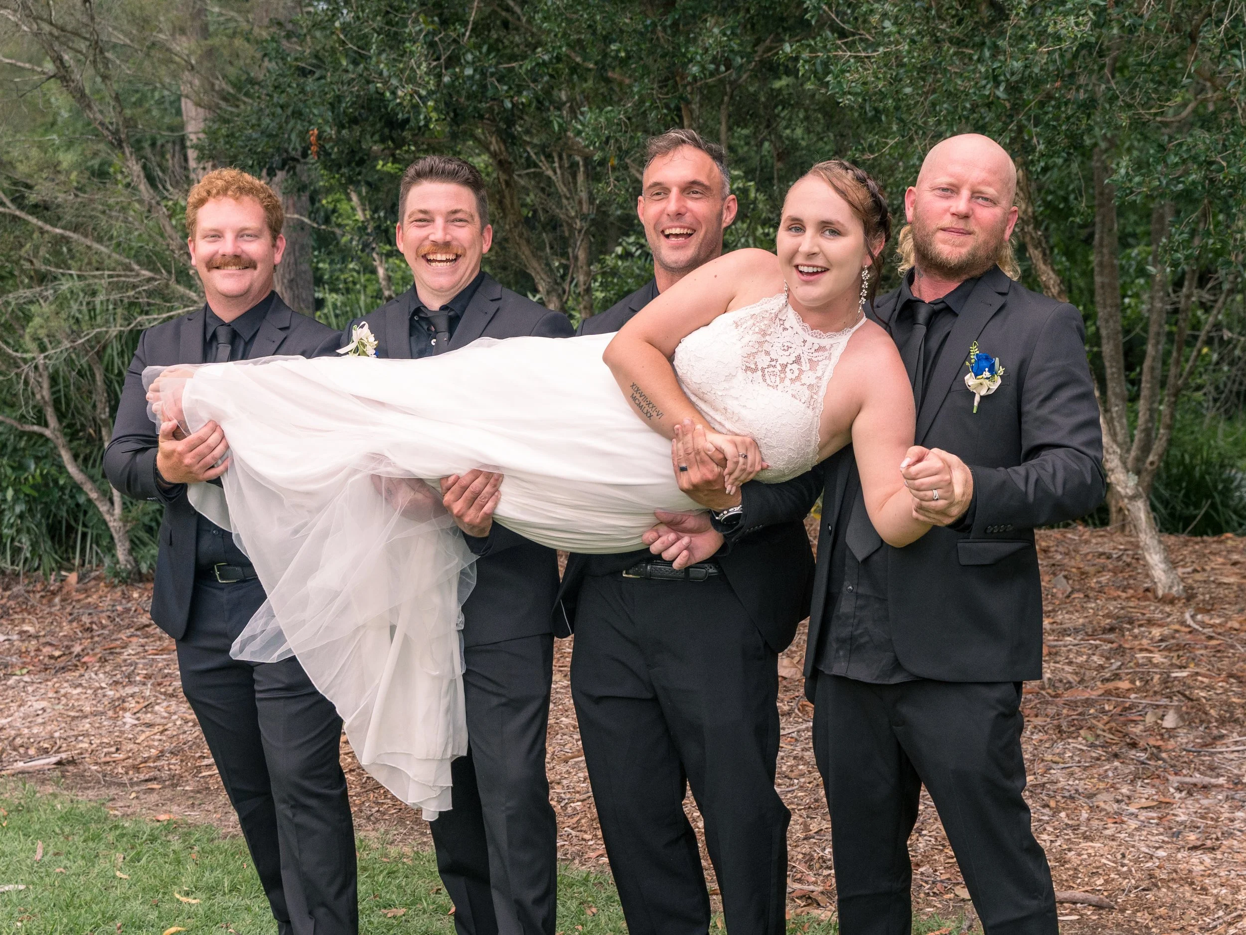 A bride in a white lace wedding dress being carried by four men in black suits outdoors with trees in the background.