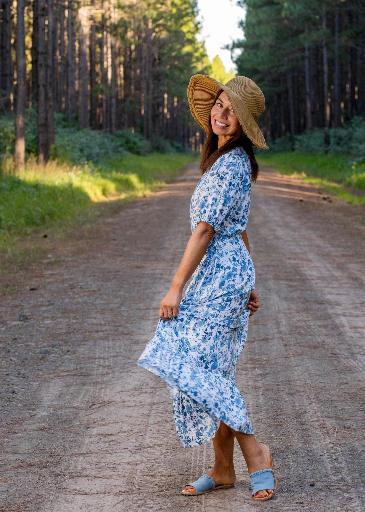 A woman in a blue and white floral dress and a wide-brimmed hat walking on a dirt path in a forest.