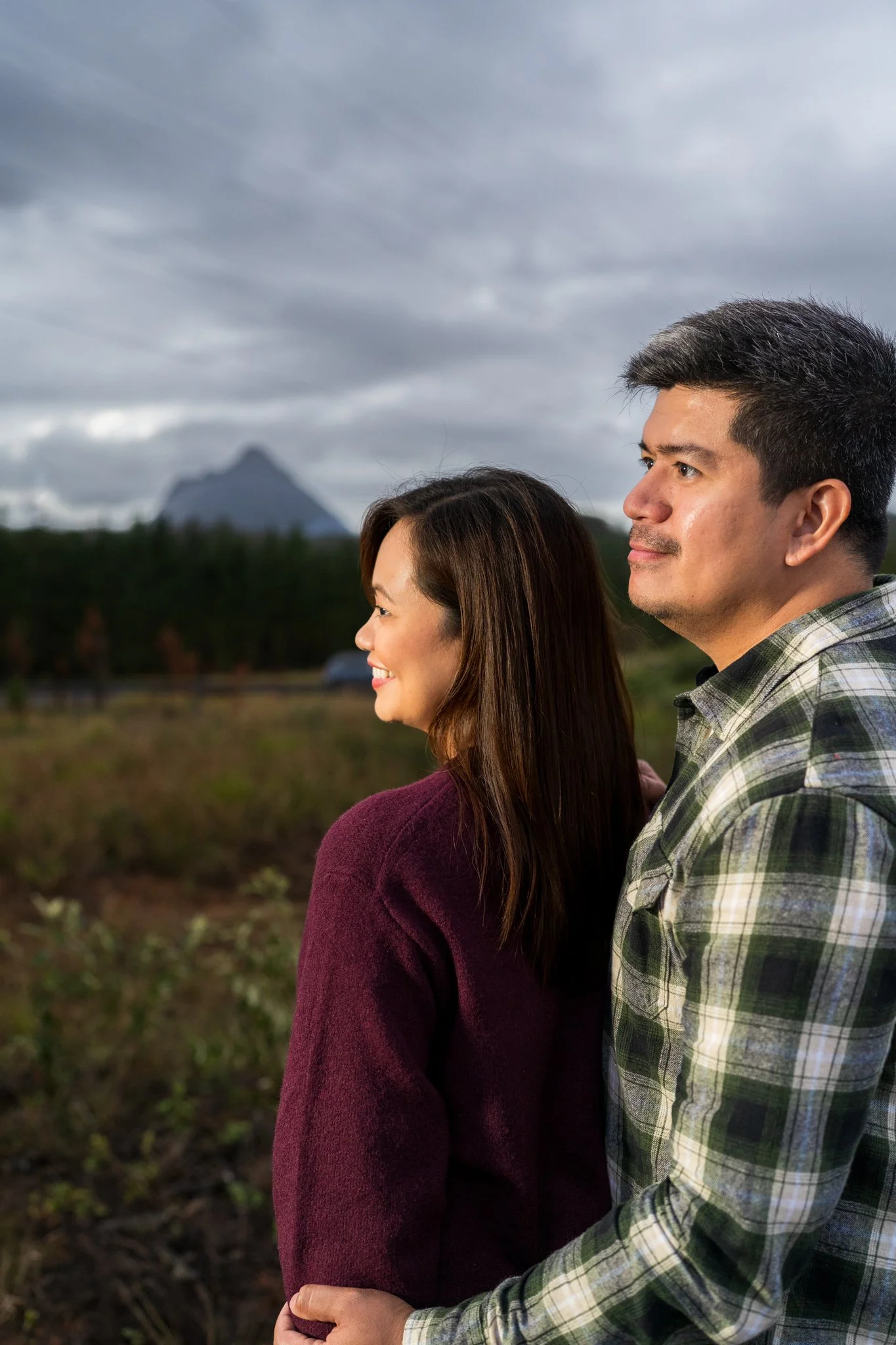 A smiling woman with long brown hair wearing a burgundy sweater and a smiling man with short dark hair and wearing a plaid shirt standing outdoors during a cloudy day with a mountain in the background.