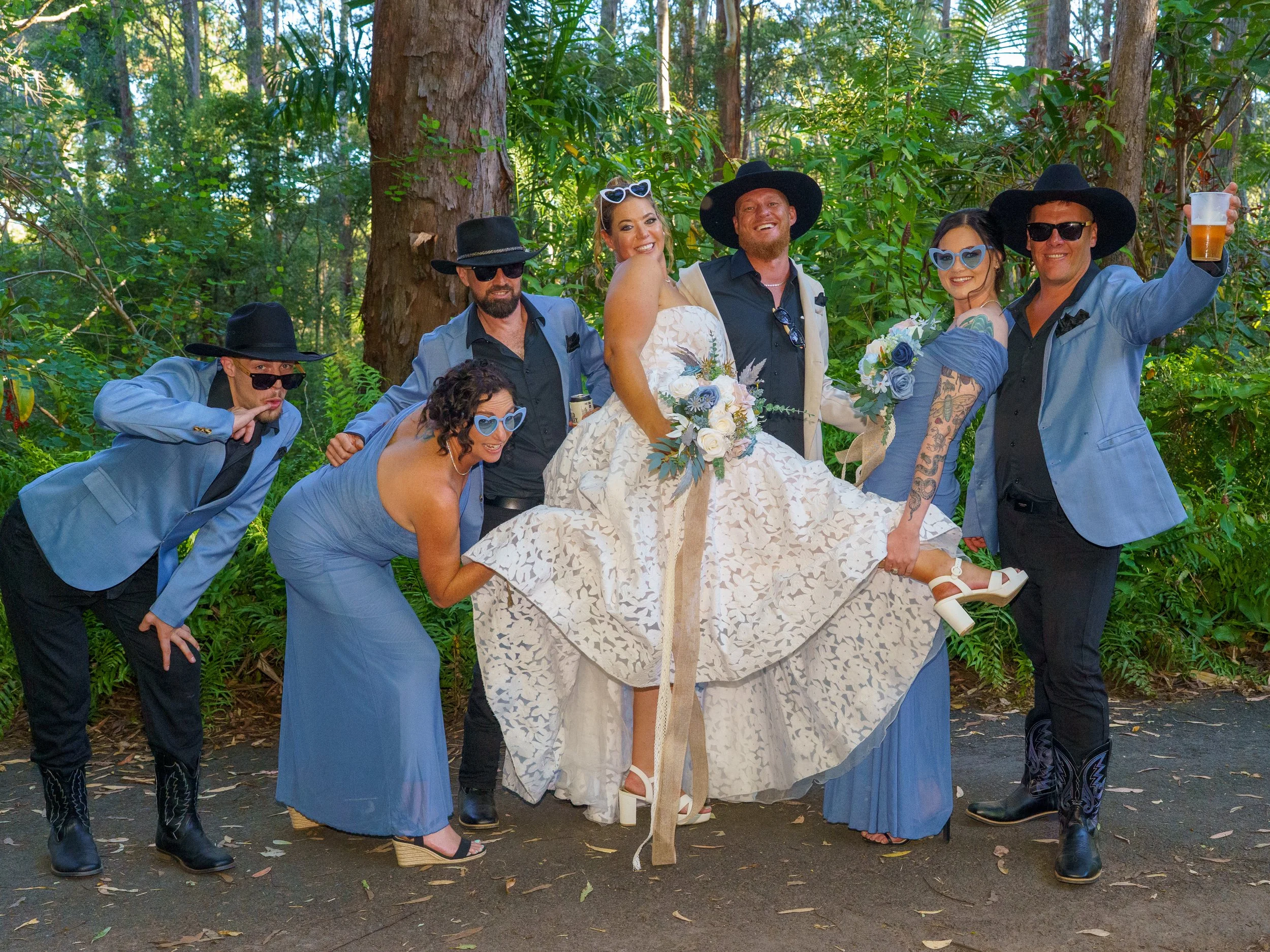 Group of people dressed in blue suits and black hats, posing outdoors in a forest setting, some wearing sunglasses, with one person holding a beer glass.