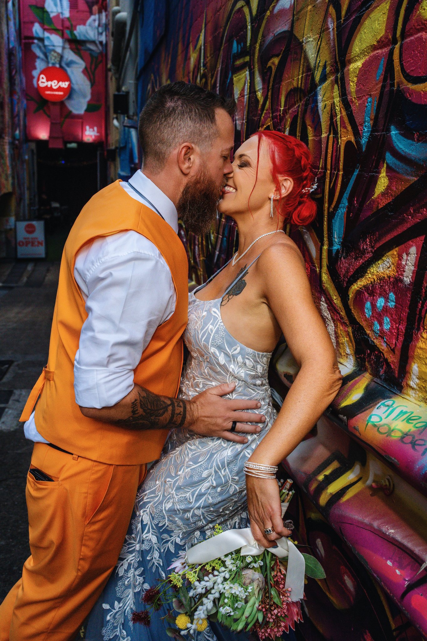 A couple standing close together in front of a colorful graffiti wall, smiling with noses touching. The man has a beard and tattoos, wearing an orange vest over a white shirt. The woman has bright red hair styled in an updo with braids, and is dressed in a patterned dress, holding a bouquet of flowers.