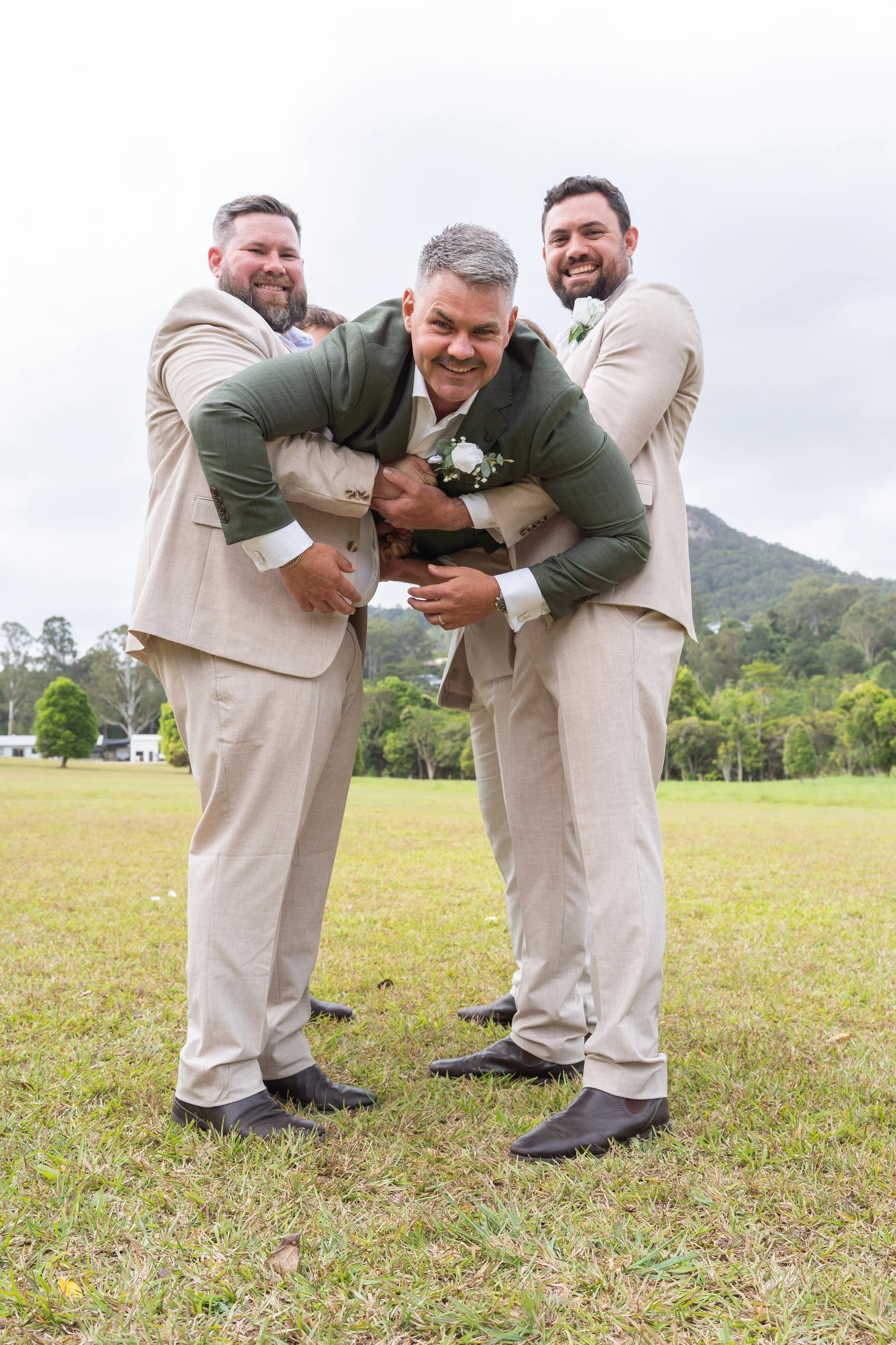 Four men in suits, standing on grass outdoors, playfully holding each other in a group hug, with a mountain and trees in the background.