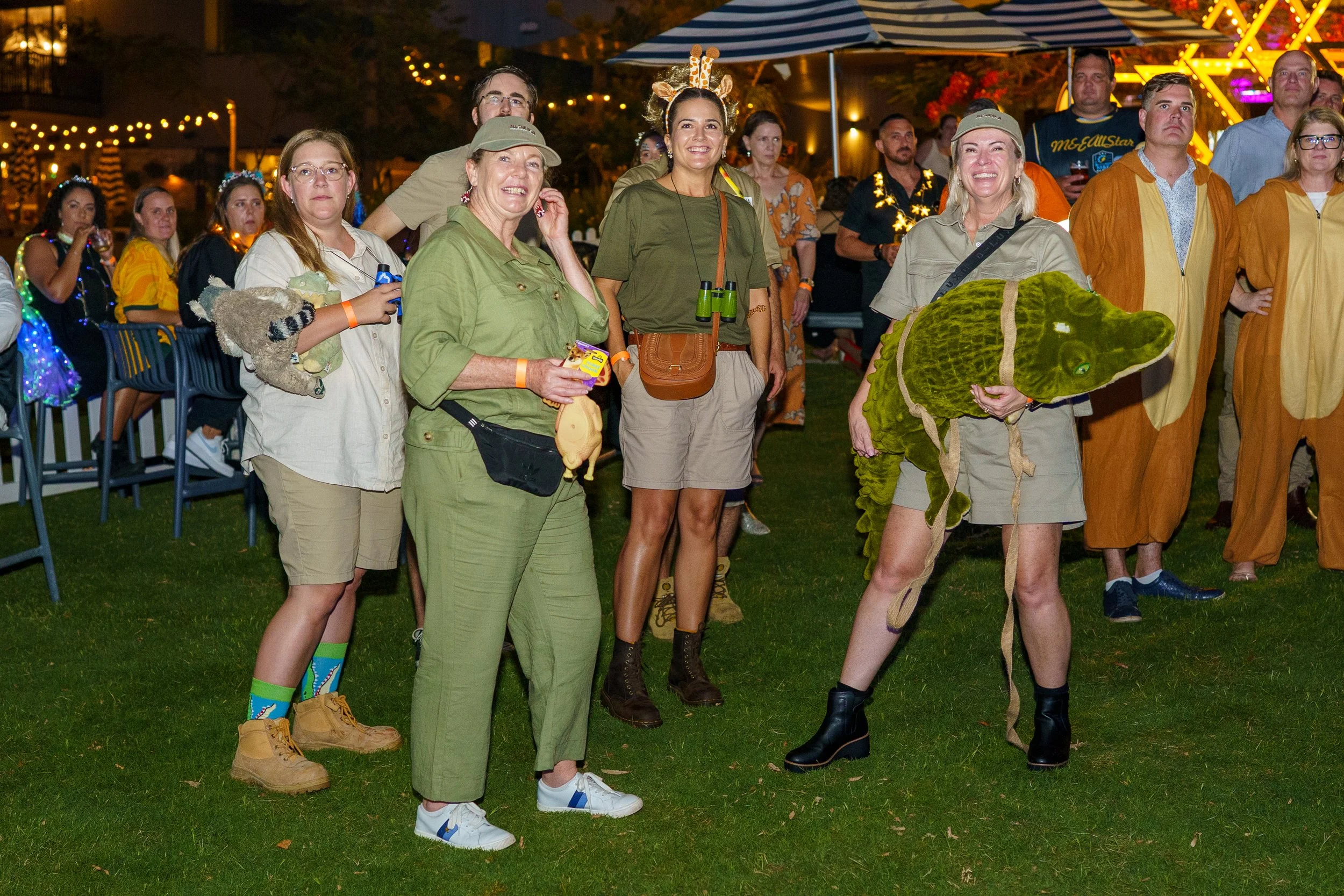 Group of people at an outdoor nighttime event, some dressed in safari or animal costumes, with festive lights and a ferris wheel in the background.