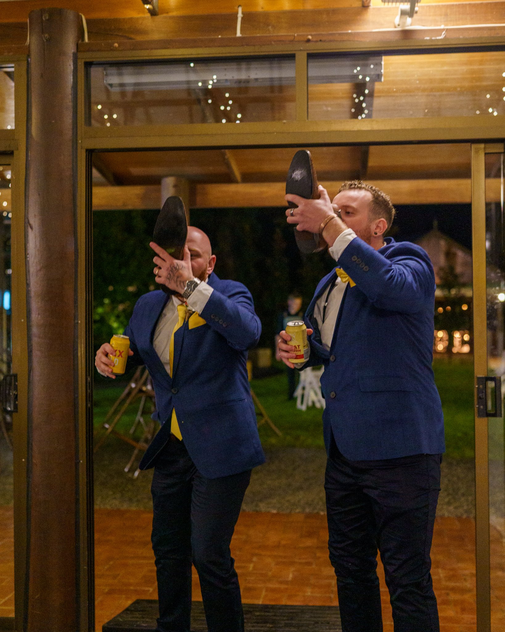Two men dressed in formal tuxedos with yellow bow ties are showcasing their shoes in front of a mirror at an indoor event. They are smiling and holding yellow cans in their hands.
