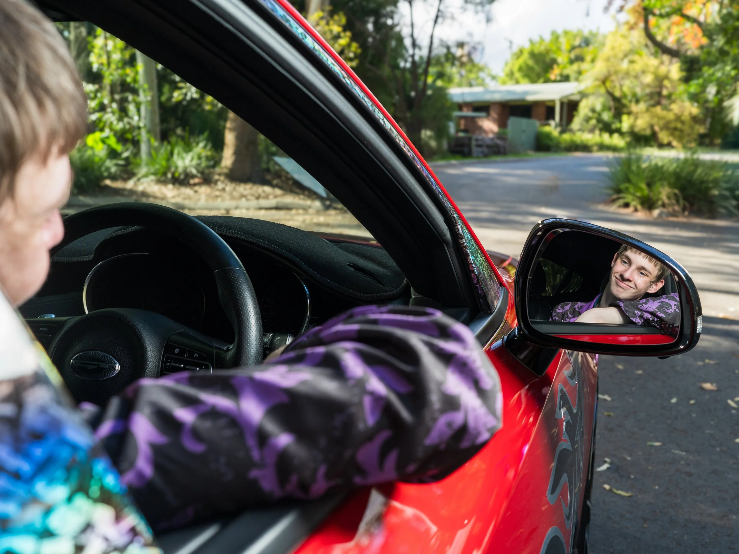 A person sitting inside a red car, looking at a reflection of a smiling man in the side mirror. The person appears to be adjusting the rearview mirror and is wearing a jacket with purple and black patterns. The background shows a suburban street with