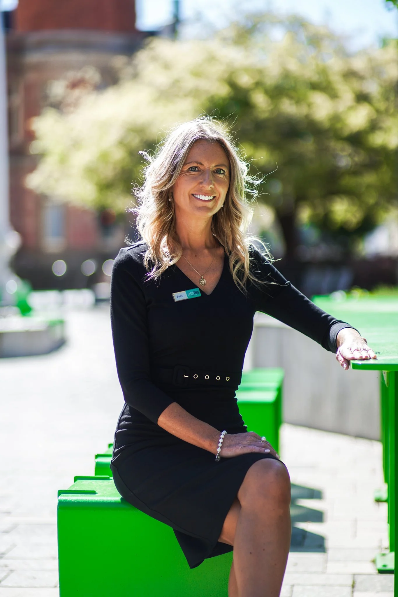 Beck has long brown blonde hair. She is sitting at a green bench outside resting her arm on a table. She is wearing a black skirt and top and is smiling warmly.