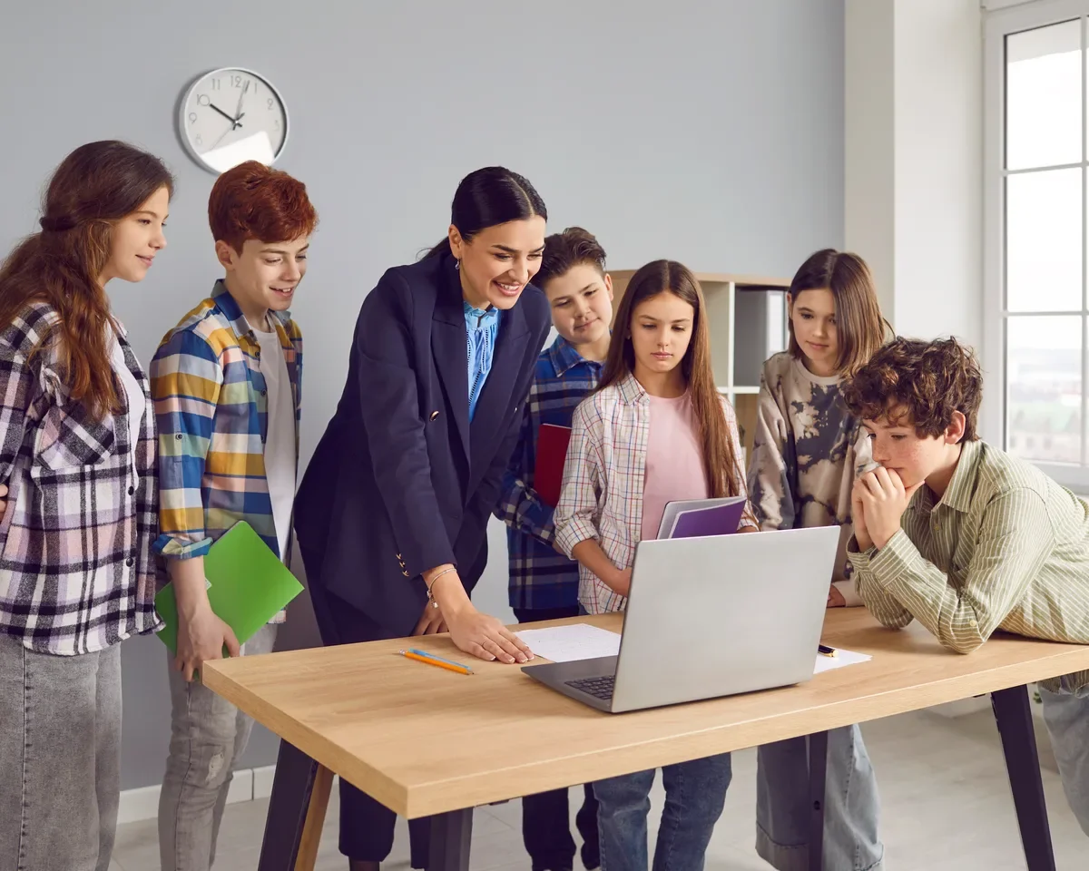 Teacher showing a laptop screen to six students gathered around a desk in a classroom.