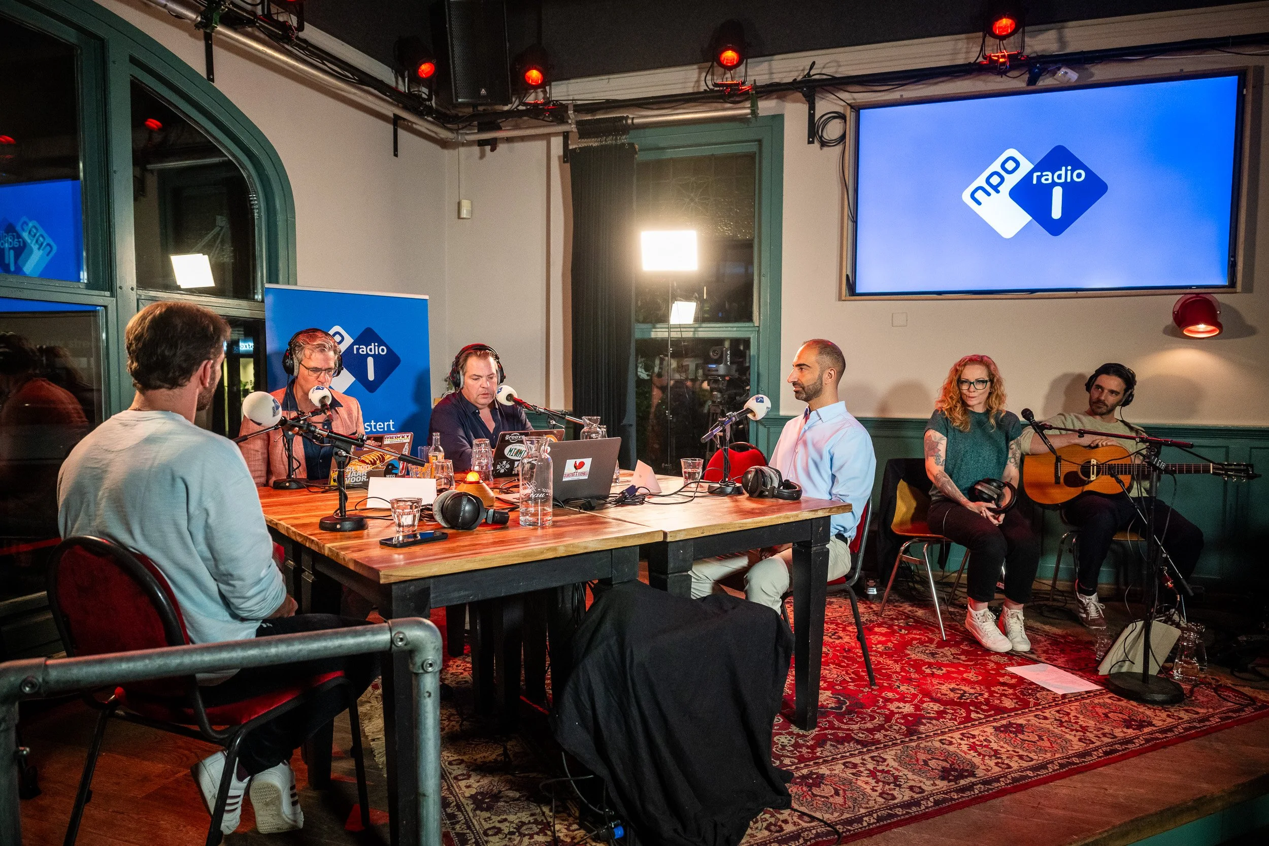 Een radiostudio met zes mensen, drie mannen en drie vrouwen, met microfoons, koptelefoons, laptops en muziekinstrumenten, en een groot scherm met het logo van NPO Radio 1.