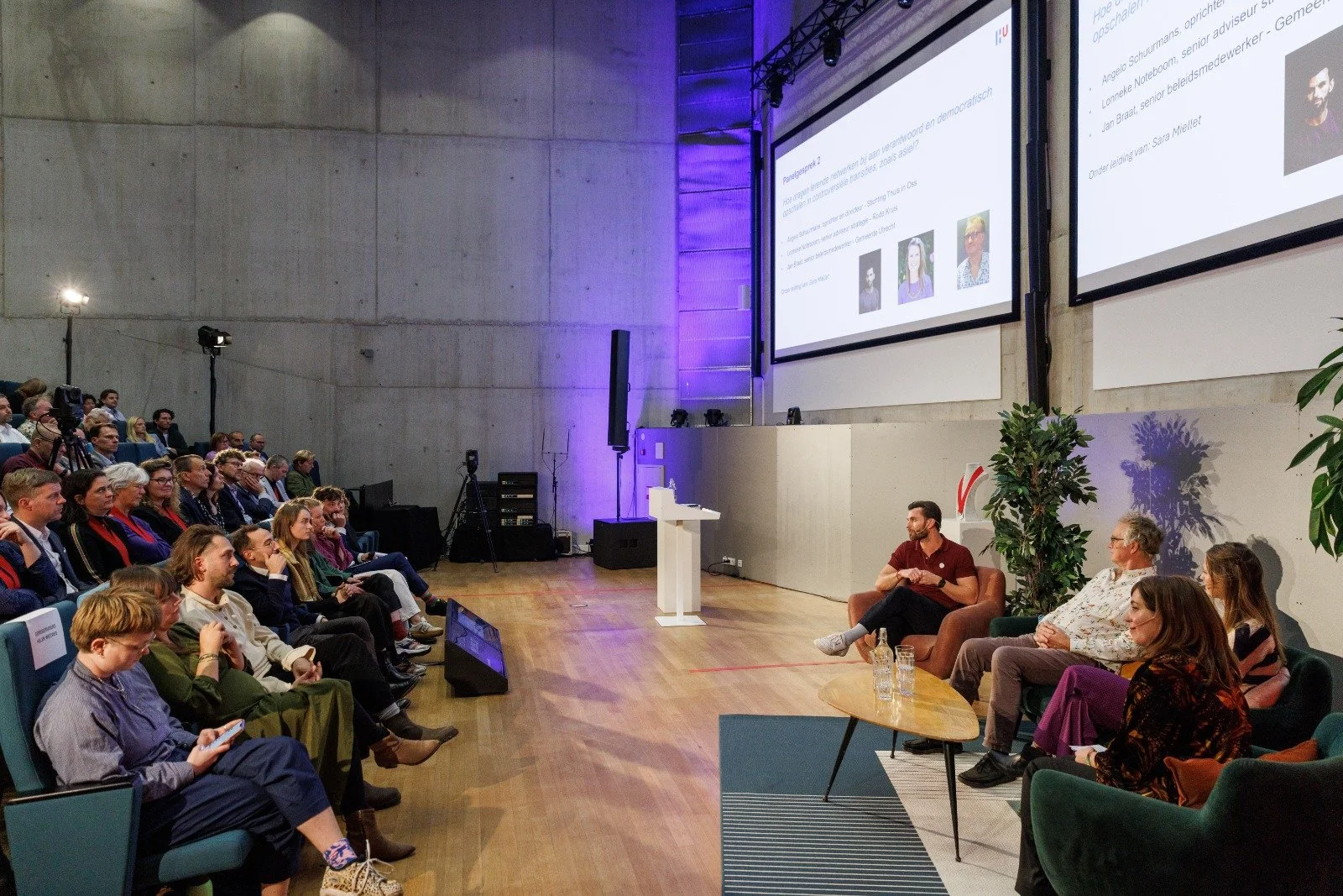 Een grote groep mensen zit in een auditorium, attent luisterend naar een presentatie. Aan de rechterkant van het beeld zitten vier mensen op een bankje, terwijl een groot scherm achter hen informatie toont. Er is een witte spreektafel met een microfoon en een paar glazen water.