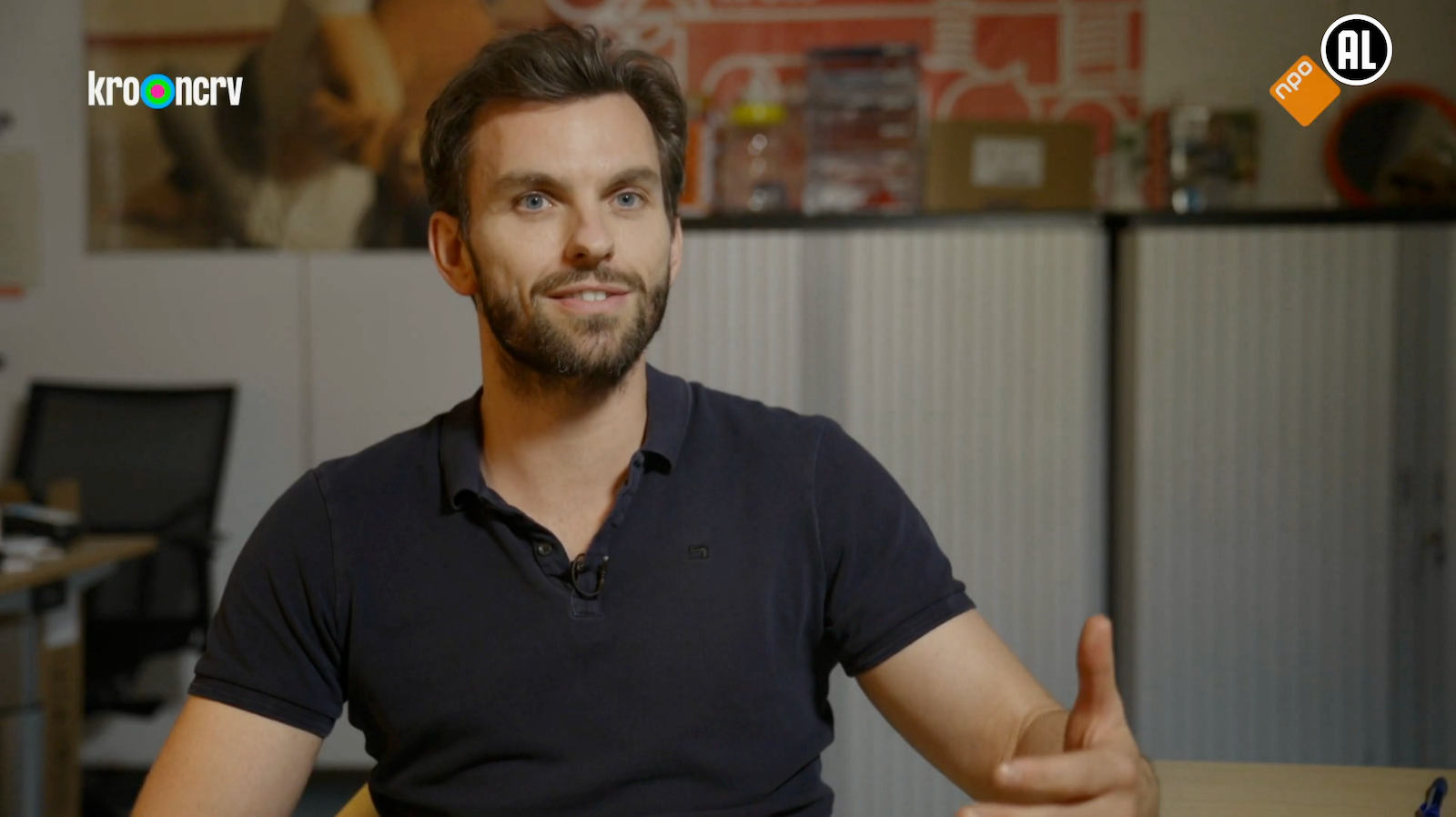 Angelo Schuurmans zit aan een tafel in een kantoor, draagt een zwart polo en spreekt in een interview.