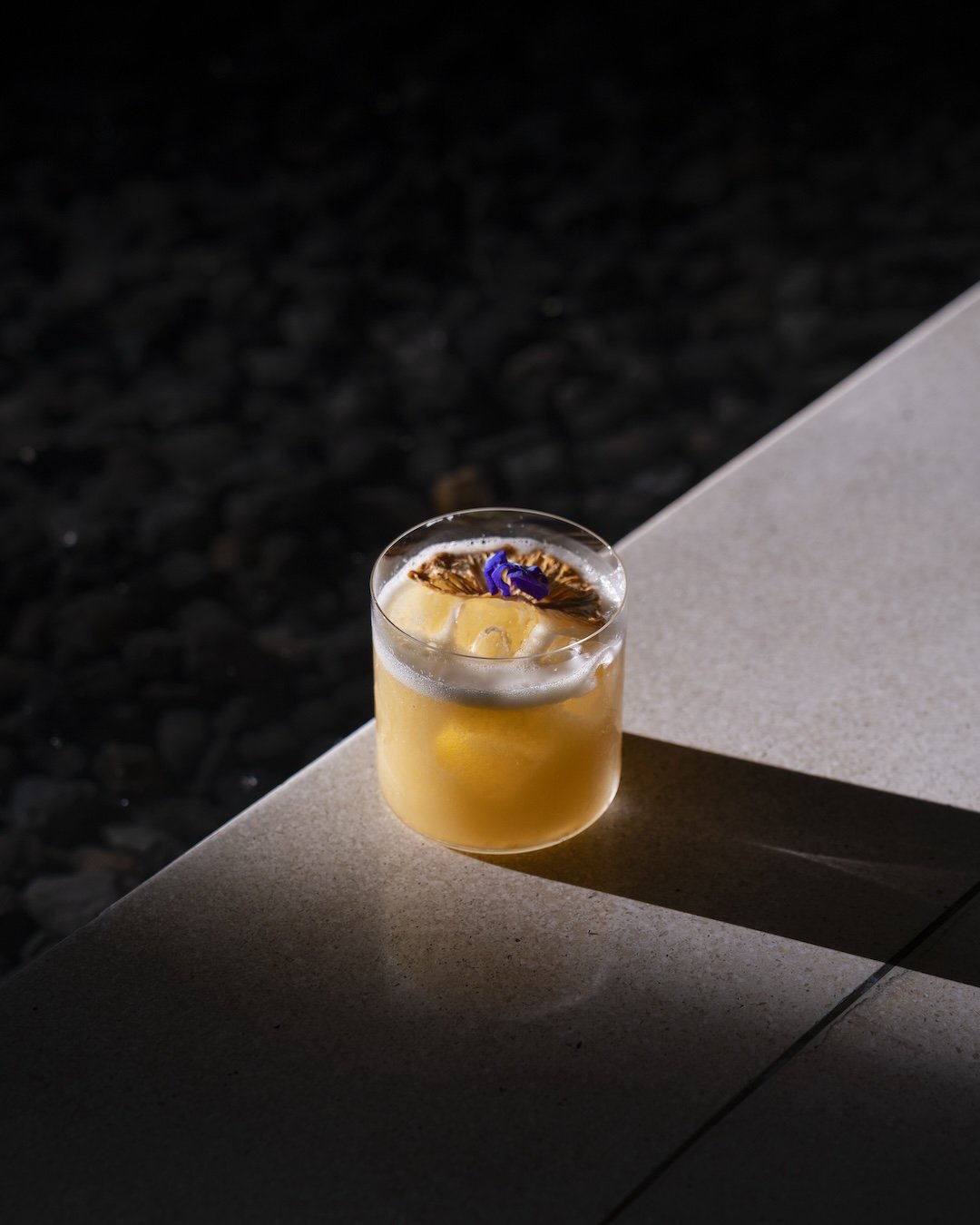 A glass with an amber cocktail garnished with a dried citrus slice and a purple flower, placed on a stone surface with a dark background.