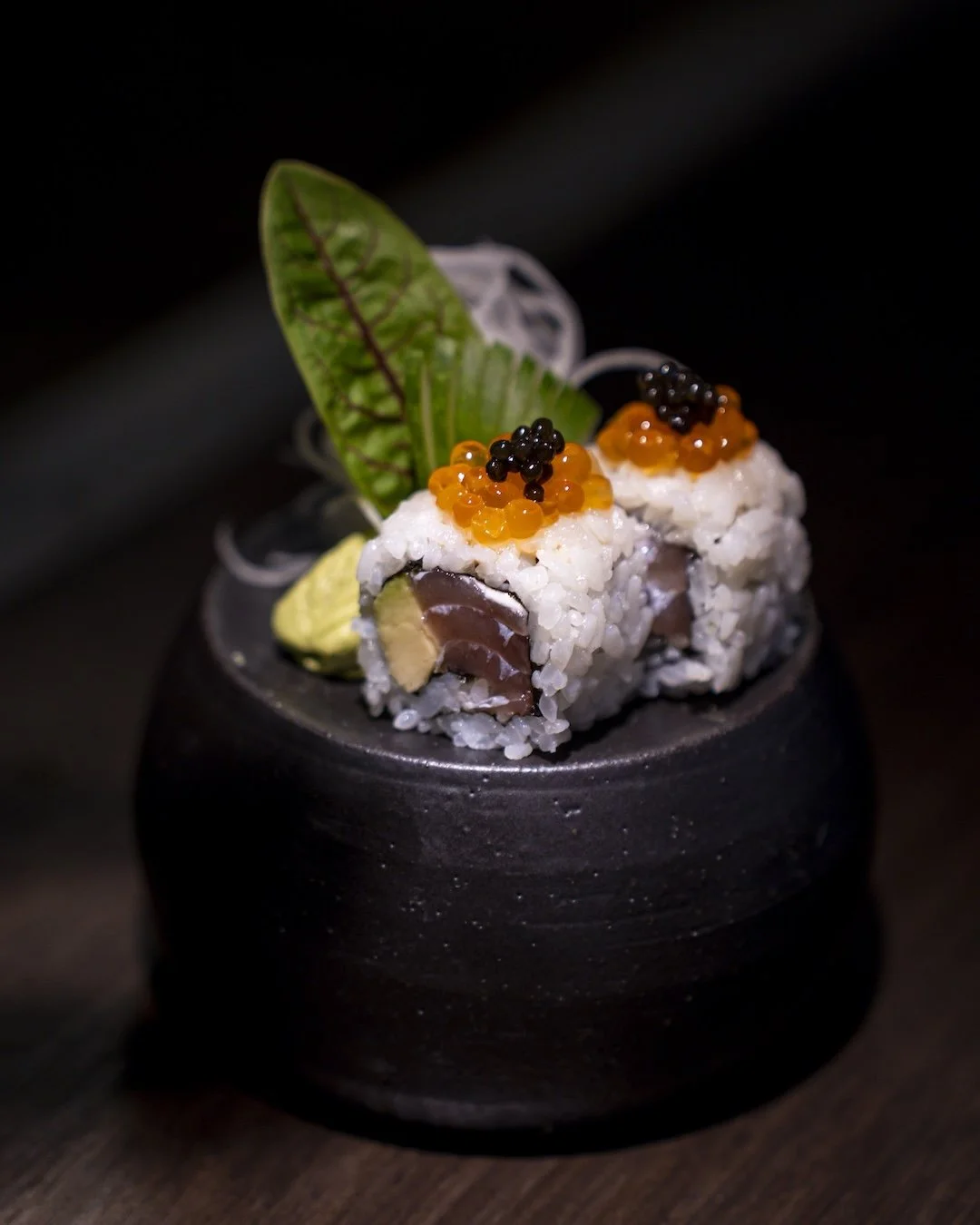 Close-up of sushi rolls topped with fish roe and caviar, garnished with a leaf and wasabi on a black plate.