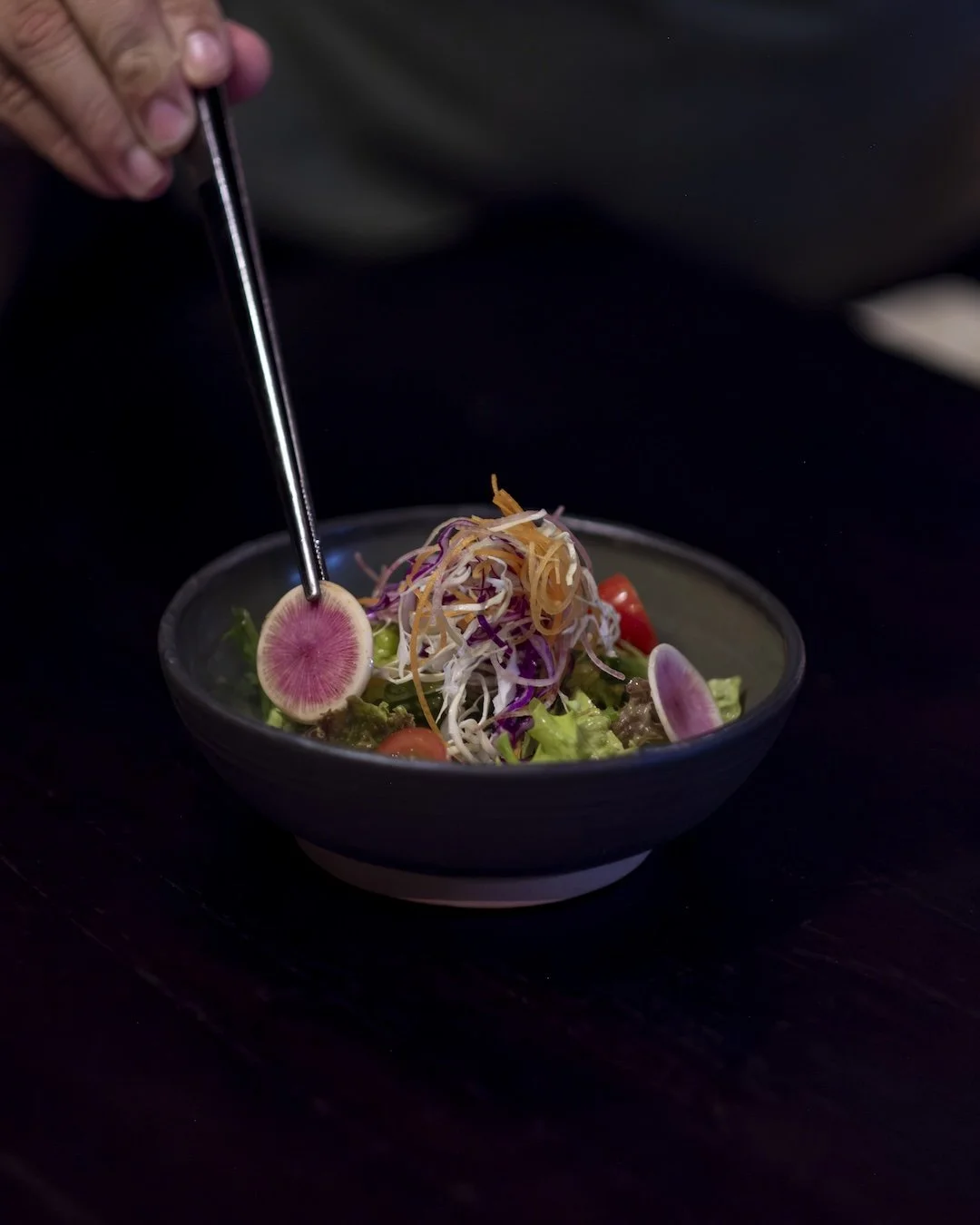 Person using tweezers to place a slice of radish on a colorful salad in a black bowl