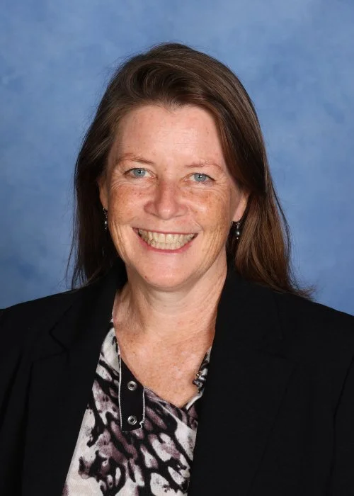 A smiling woman with short gray hair, glasses, pearl earrings, wearing a black floral jacket, an orange scarf, and a gold necklace.