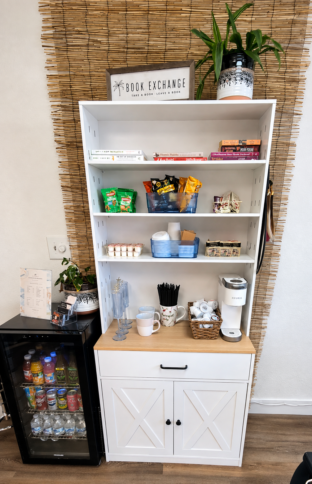 A white shelf with food and drinks setup, a small black mini fridge filled with drinks, and a bamboo wall background. The shelf contains coffee cups, a coffee maker, snacks, disposable cups, and a small plant, with a framed sign on top that reads 'Book Exchange: Take a Book, Leave a Book.'
