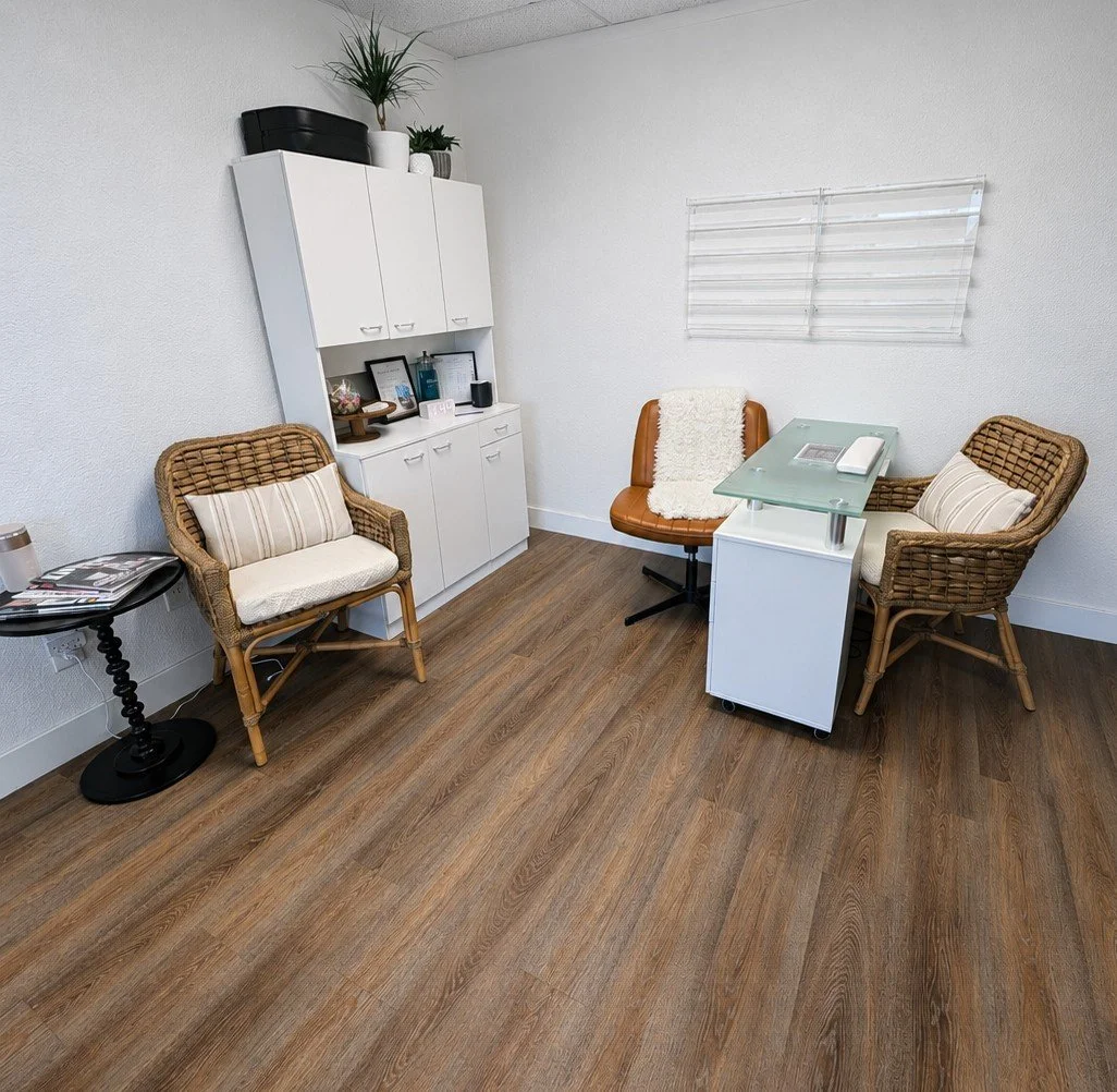 A small medical or therapy office with a white cabinet, two rattan chairs with cushions, a brown leather chair with a throw blanket, a glass-top desk with a monitor, and a small black side table with magazines. The room has wood flooring and white walls.
