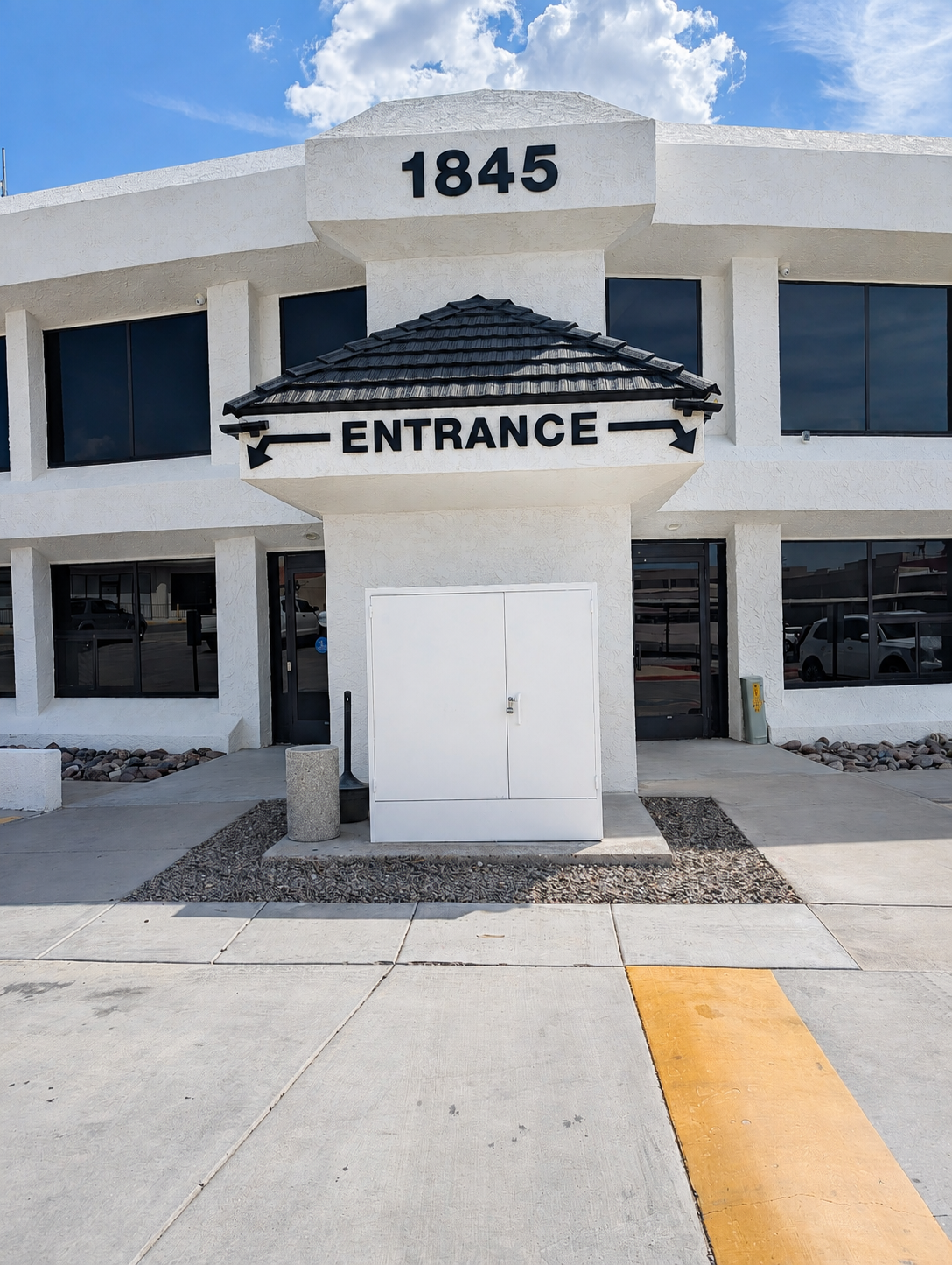 White commercial building with the number 1845 on top, a small roofed sign indicating entrance with arrows pointing left and right, large windows, and a sidewalk with yellow and gray markings.