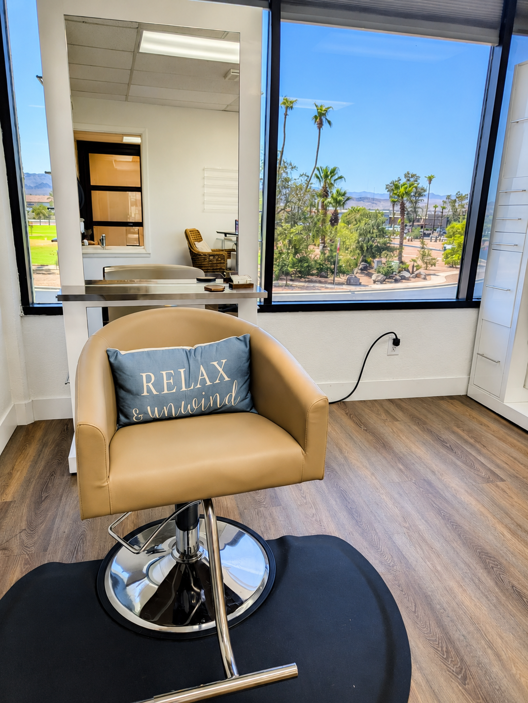 Salon chair with a blue pillow that says 'RELAX & UNWIND' in a hair salon with a window showing palm trees and a sunny outdoor scene.