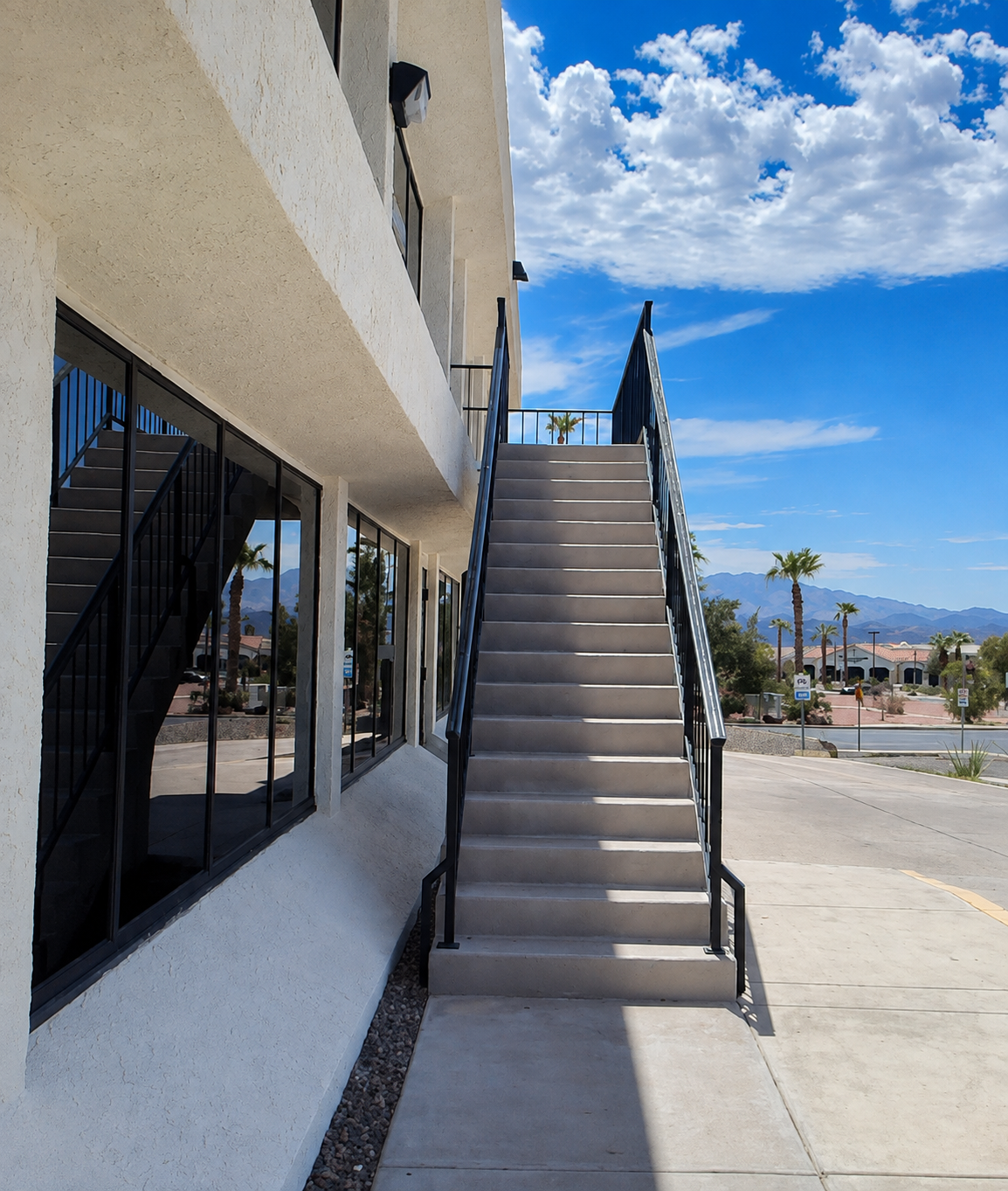 Concrete staircase outside a white building with large windows, leading to an upper level, with a mountain and palm trees in the background under a partly cloudy blue sky.