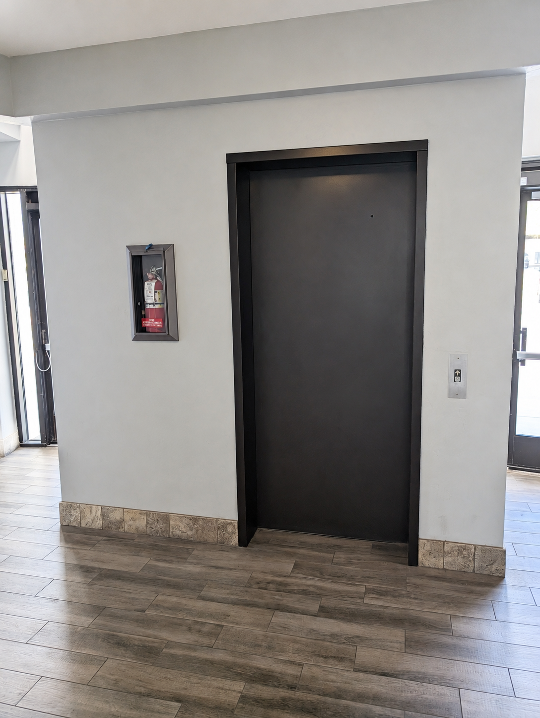 Black elevator door on a white wall with fire extinguisher inside a glass case on the left and elevator button on the right.