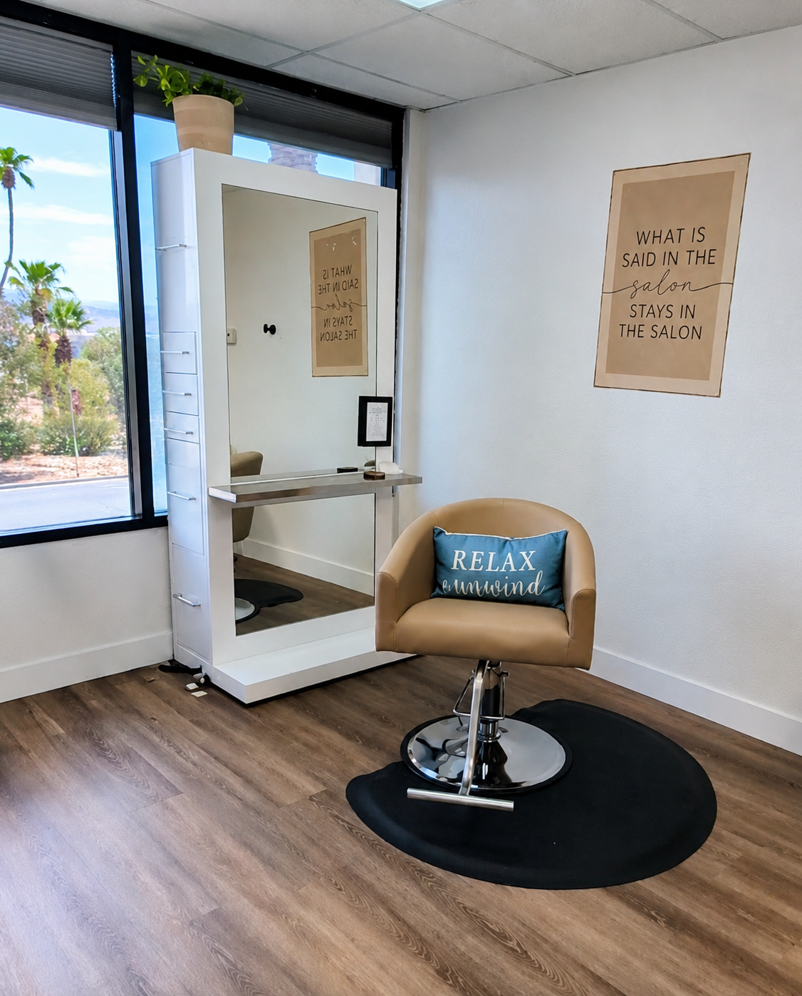 Salon chair with blue pillow that reads "RELAX and unwind" placed on a black rug, nearby a white mirror with a small counter, and a wall sign that says "What is said in the salon stays in the salon" in a salon with wooden flooring and a large window showing outdoor trees and palm trees.