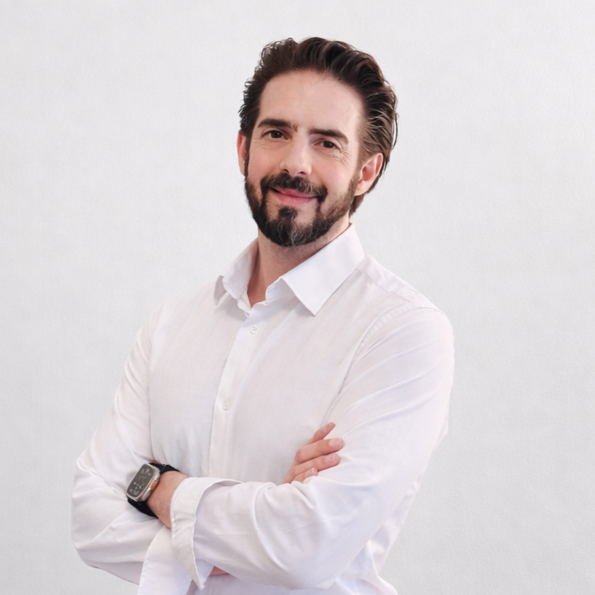 Un homme avec une barbe et des cheveux bruns, portant une chemise blanche, souriant et croisant les bras, se tenant contre un mur blanc.