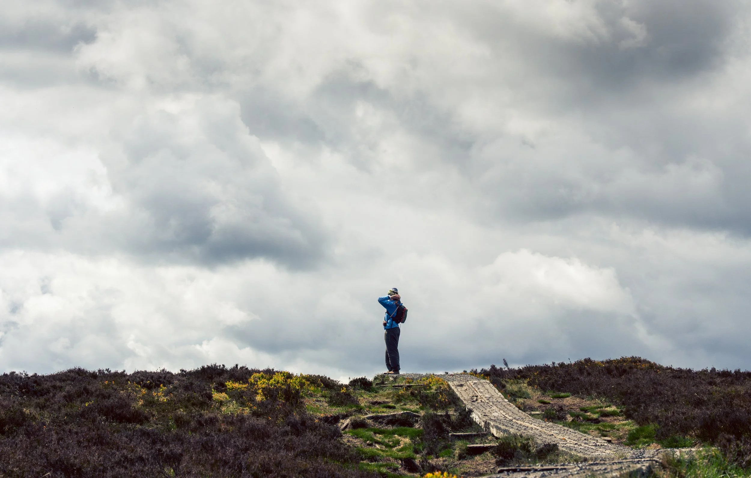 A person standing on a trail in a natural landscape with cloudy sky overhead, wearing outdoor gear and carrying a backpack.