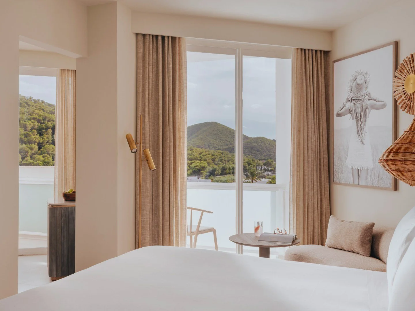 Hotel room with modern decor, featuring a large bed, beige curtains, and a balcony with mountain view. There's a sitting area with a small table and a painting on the wall.