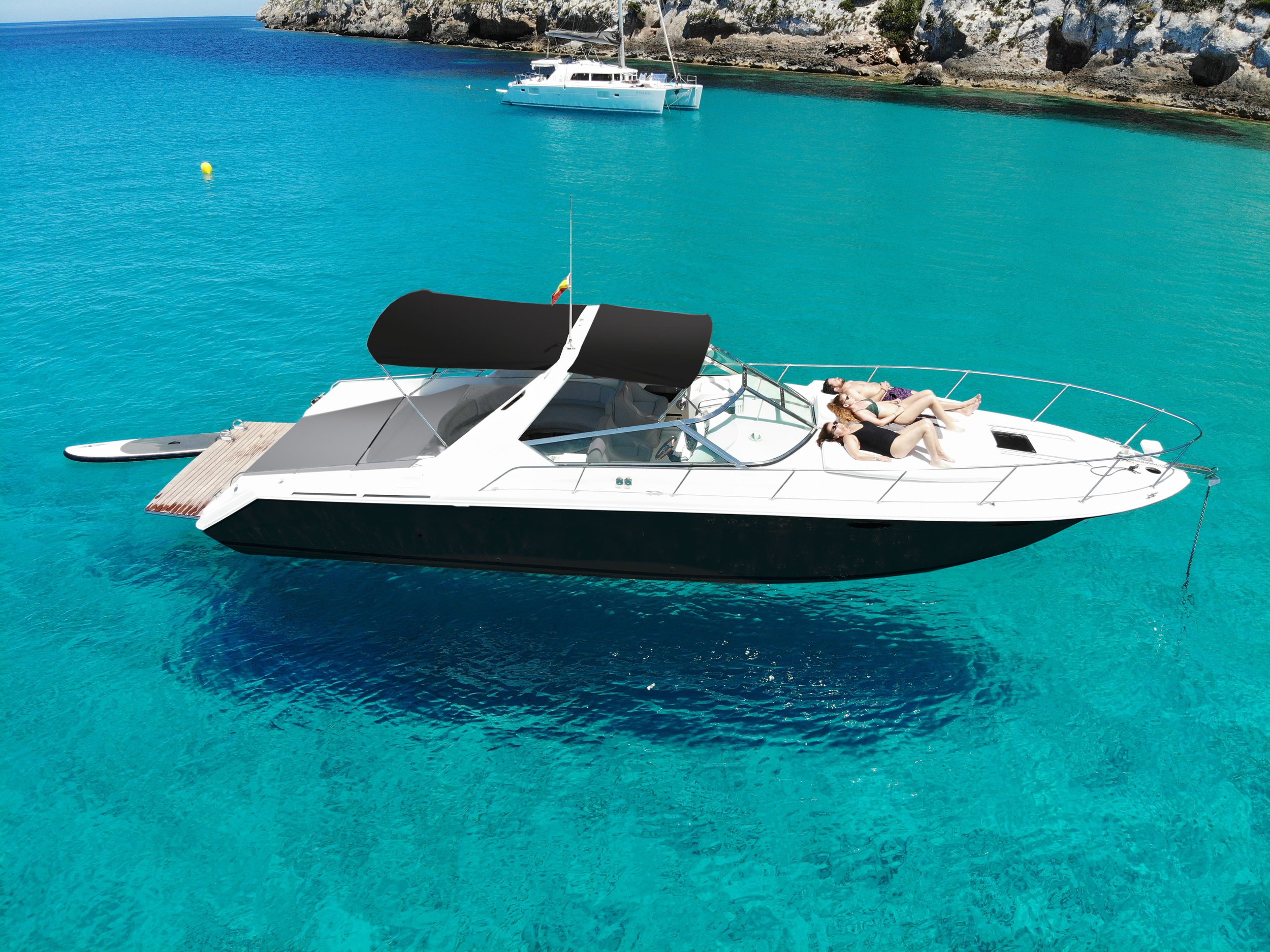 Luxury yacht on crystal clear turquoise water with people sunbathing on deck, another boat in the background near rocky shoreline.
