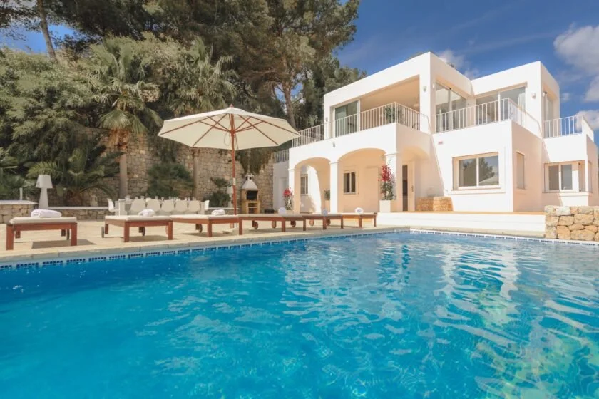 Modern white villa with a swimming pool, surrounded by trees, featuring a spacious patio with umbrellas and lounge chairs, under a clear blue sky.