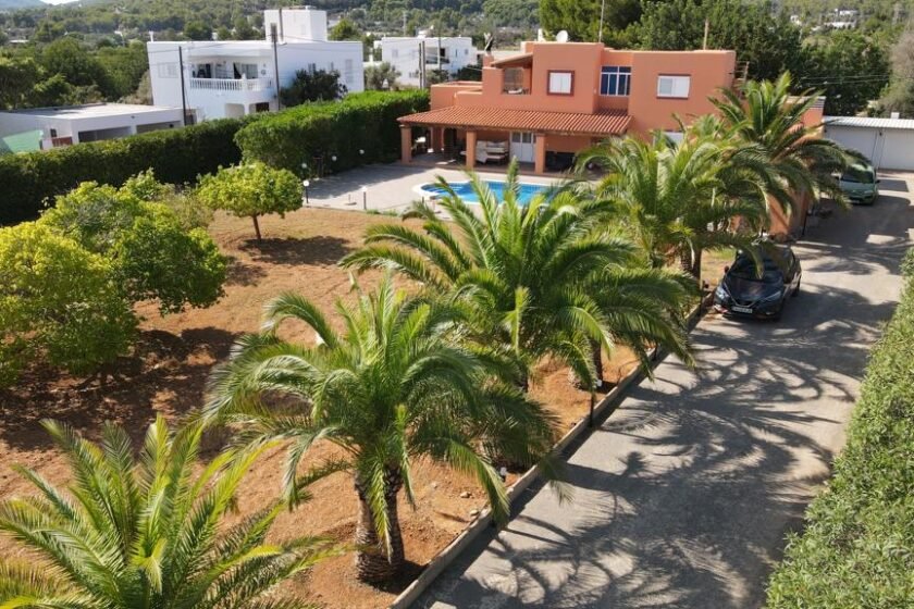 Aerial view of a house with orange exterior, swimming pool, palm trees, driveway, and surrounding greenery.