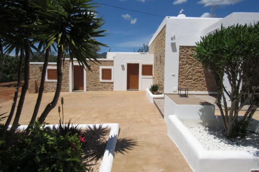 Mediterranean-style house with stone and white stucco walls, surrounded by a garden with palm trees and plants, under a clear blue sky.
