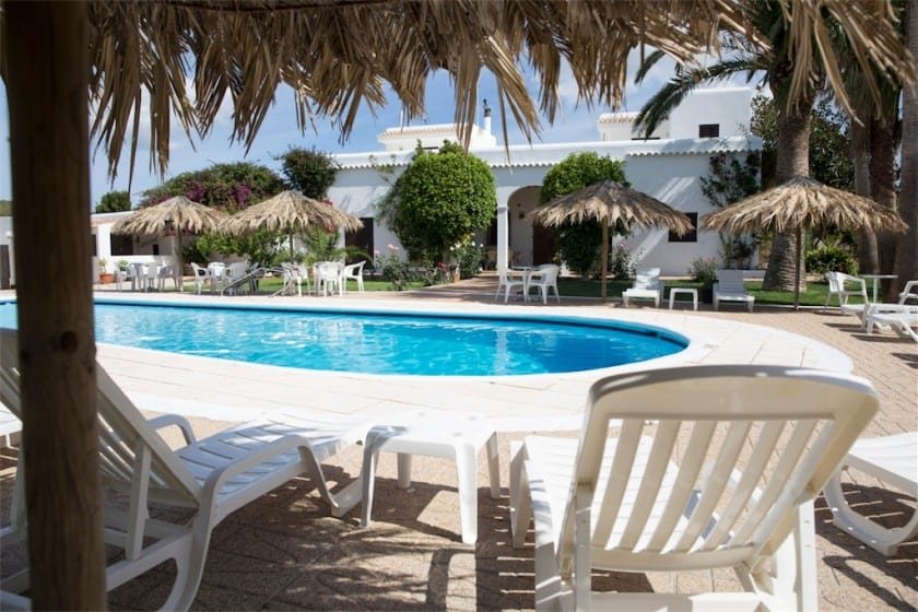 Outdoor swimming pool area with sun loungers, thatched umbrellas, and a garden in the background.