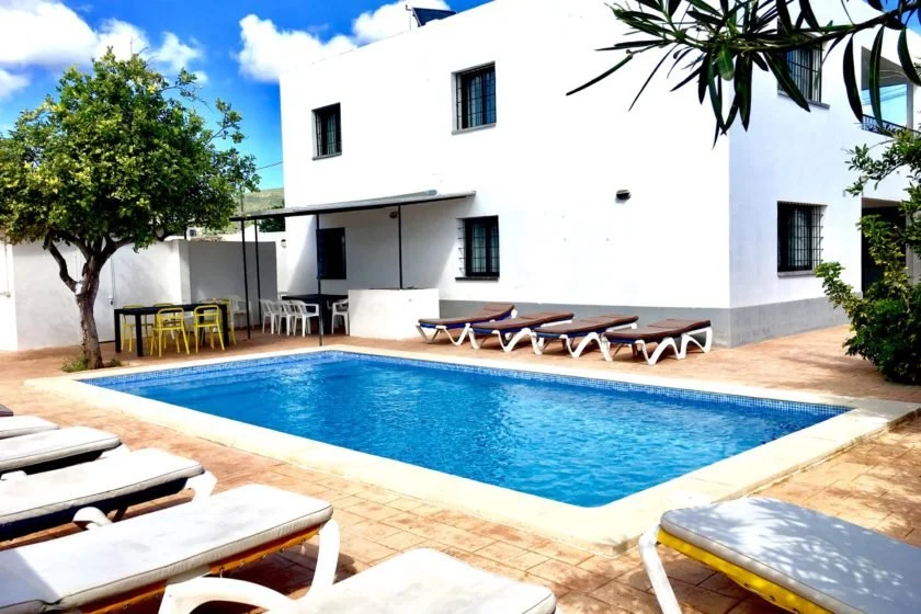 White villa with swimming pool and sun loungers, surrounded by trees, featuring outdoor seating area and bright blue sky.
