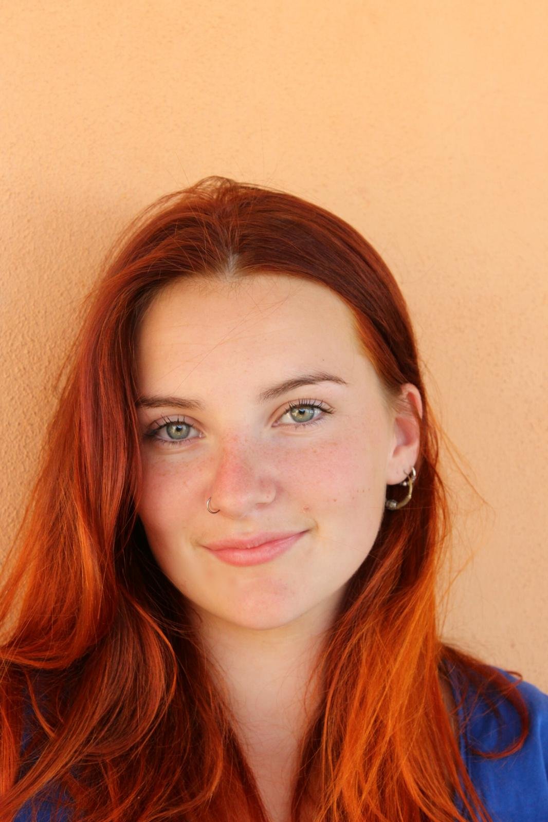 Person with red hair, nose ring, and earrings against a peach background.